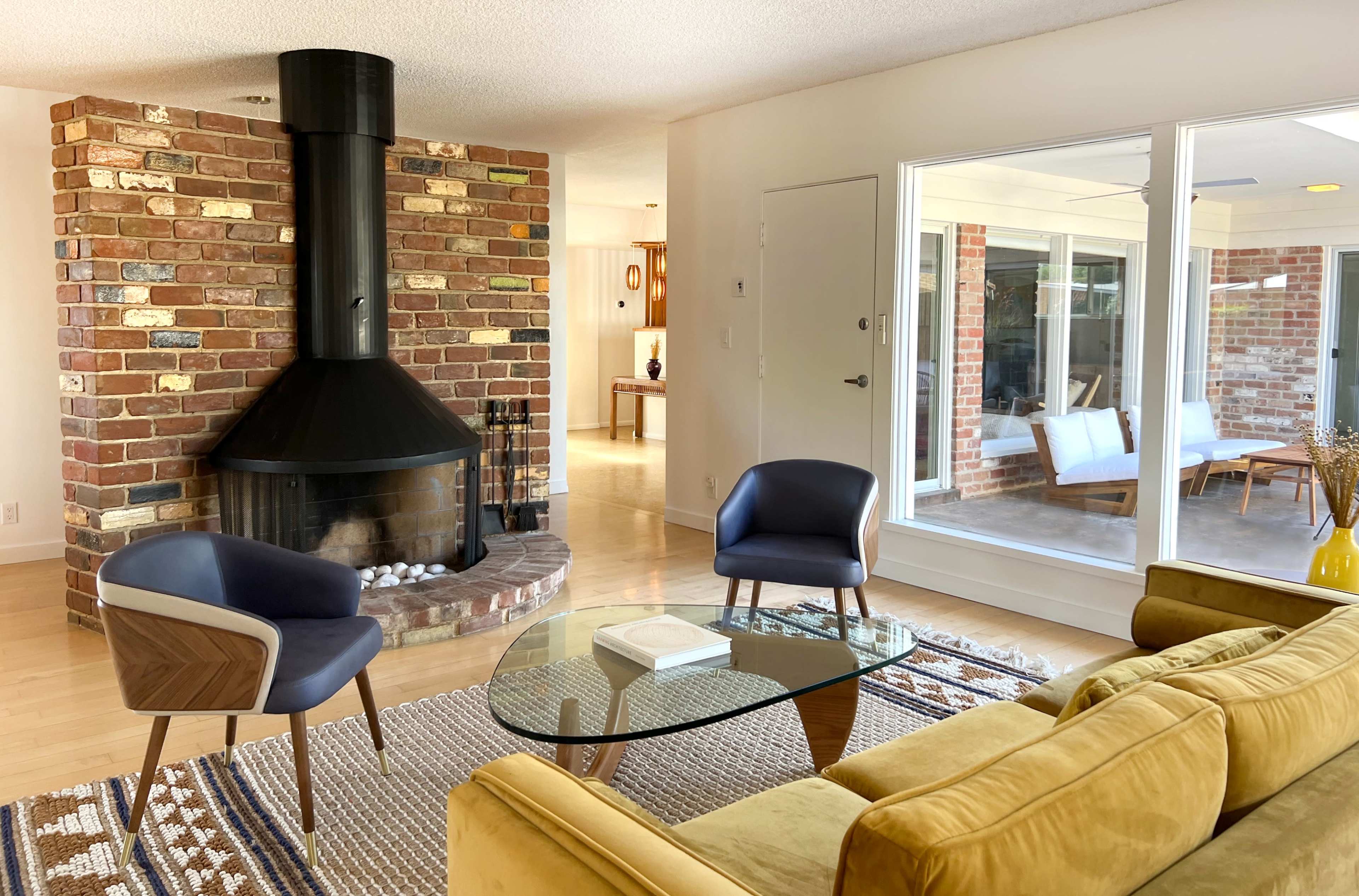 A stylish living room with a black chimney-style fireplace, two blue armchairs, a glass coffee table, and a yellow sofa, surrounded by brick walls and large windows.