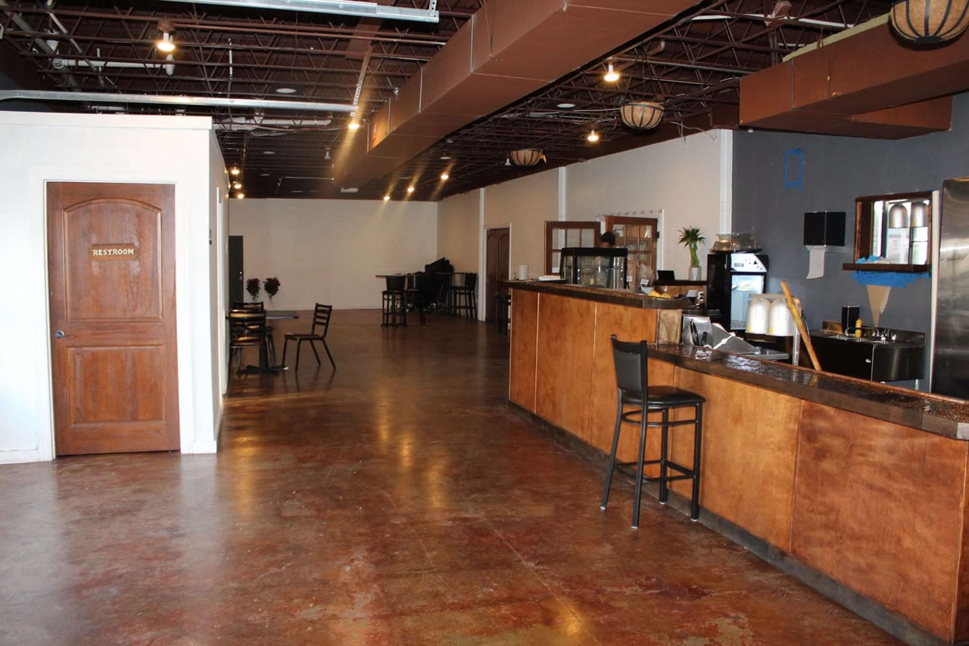 The image shows an interior of a spacious café or restaurant with a wooden counter, a few tables, and a restroom door.