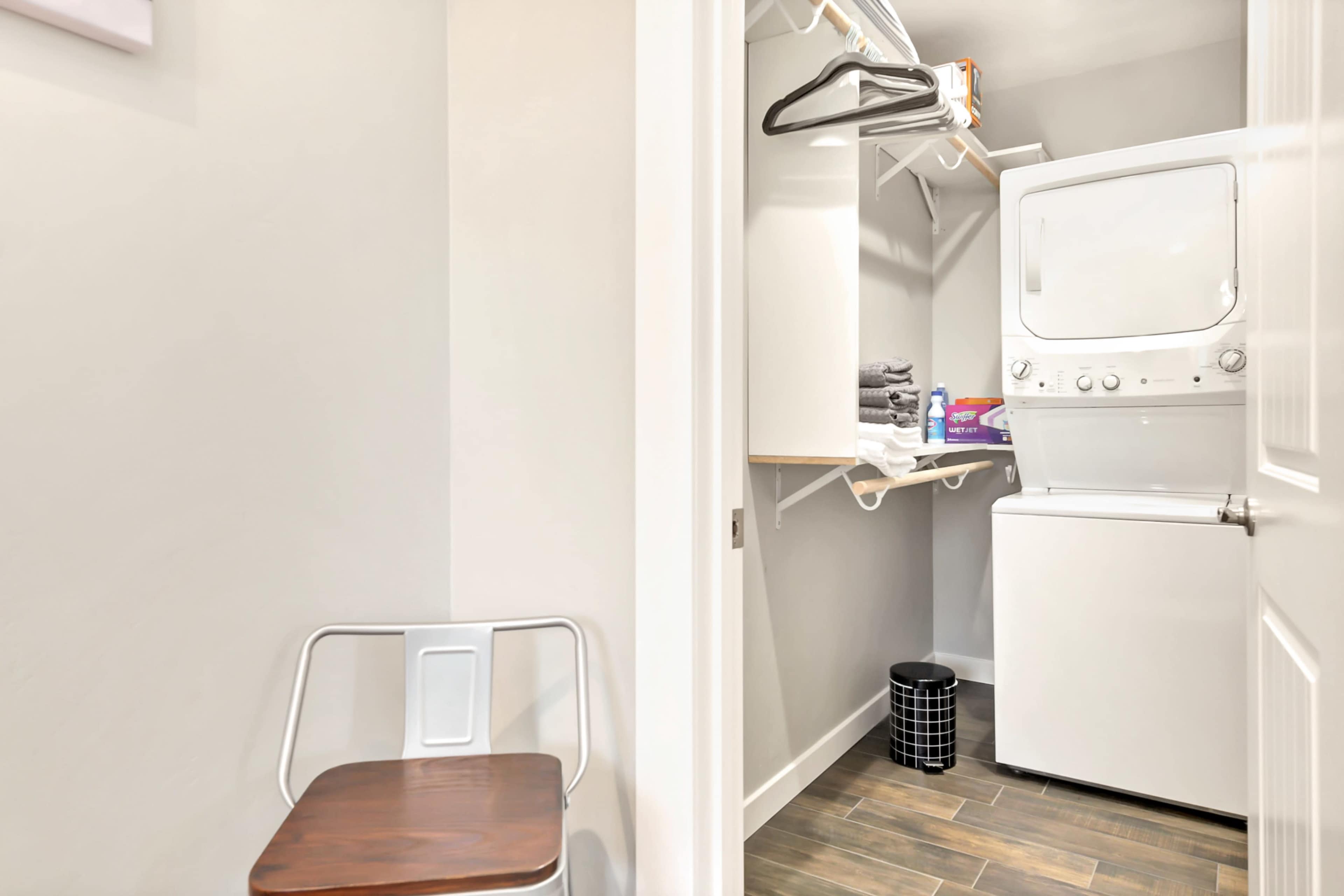 A compact laundry area features a stacked washer and dryer, shelving with towels, and a chair nearby.