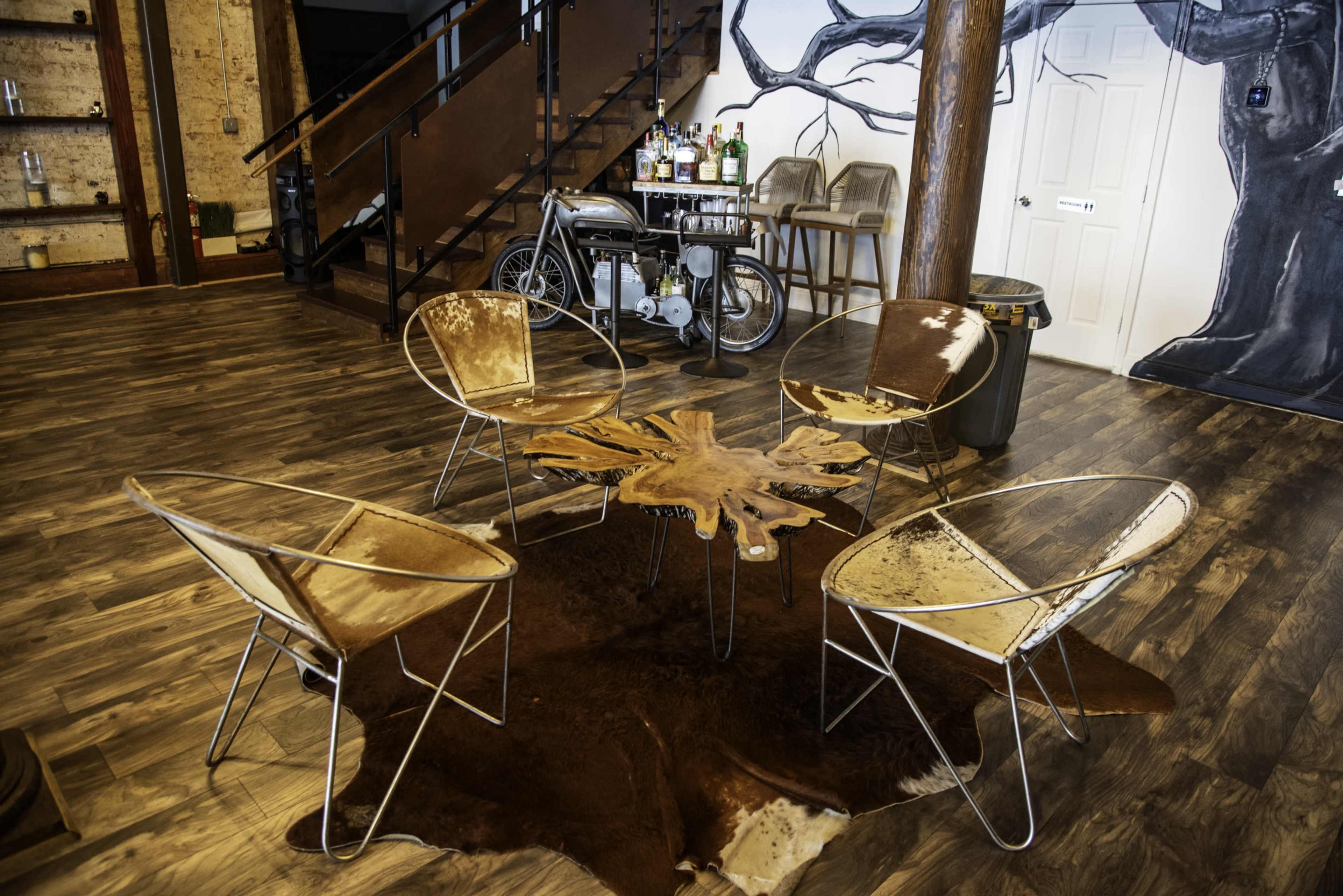 The image shows a seating area featuring four distinctive chairs around a unique table made from a tree stump, set against a backdrop of a rustic space with wooden flooring and a vintage motorcycle.