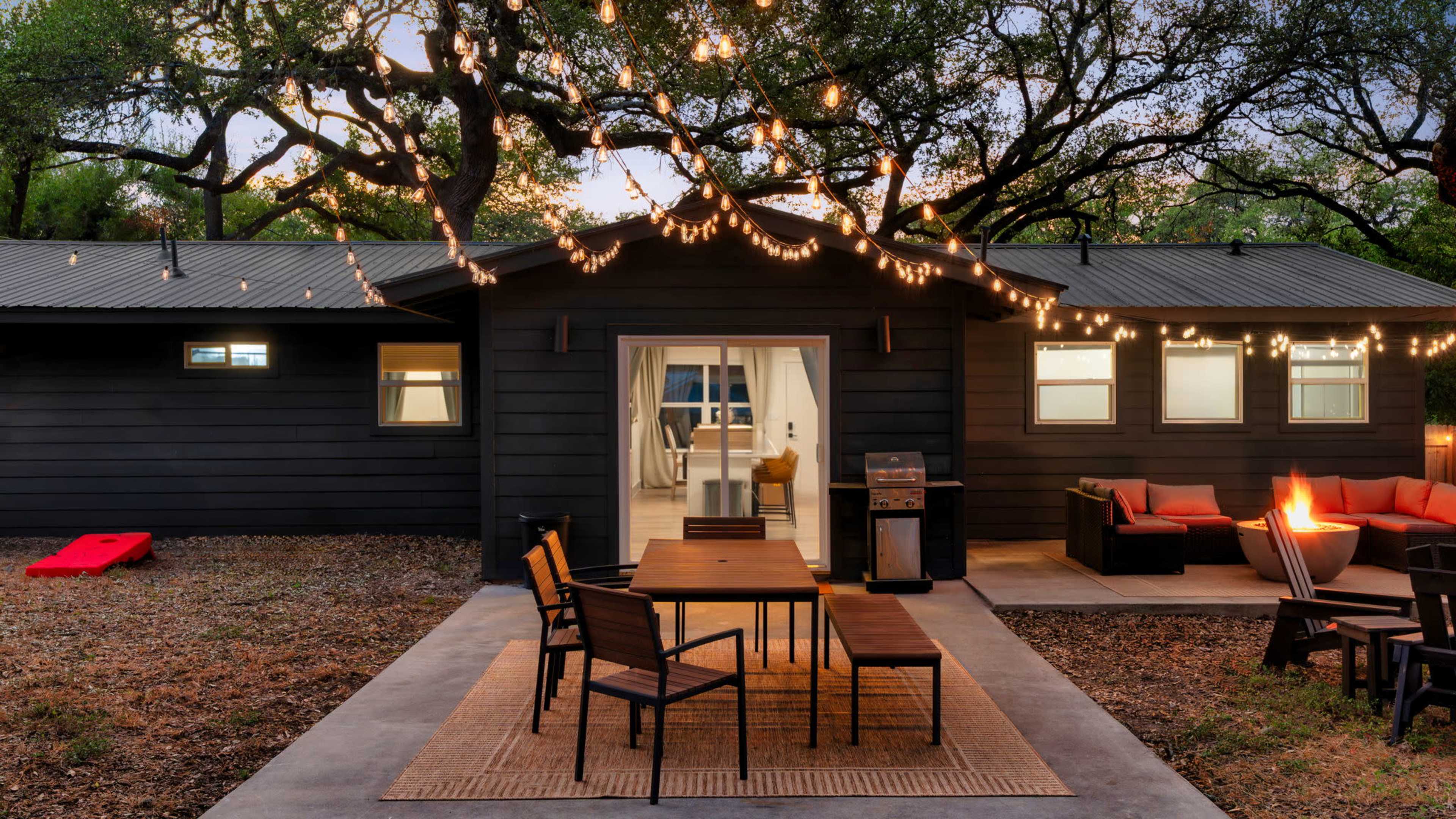 A modern black house with string lights hangs over an outdoor dining area featuring a wooden table and chairs, adjacent to a cozy seating area with a fire pit.