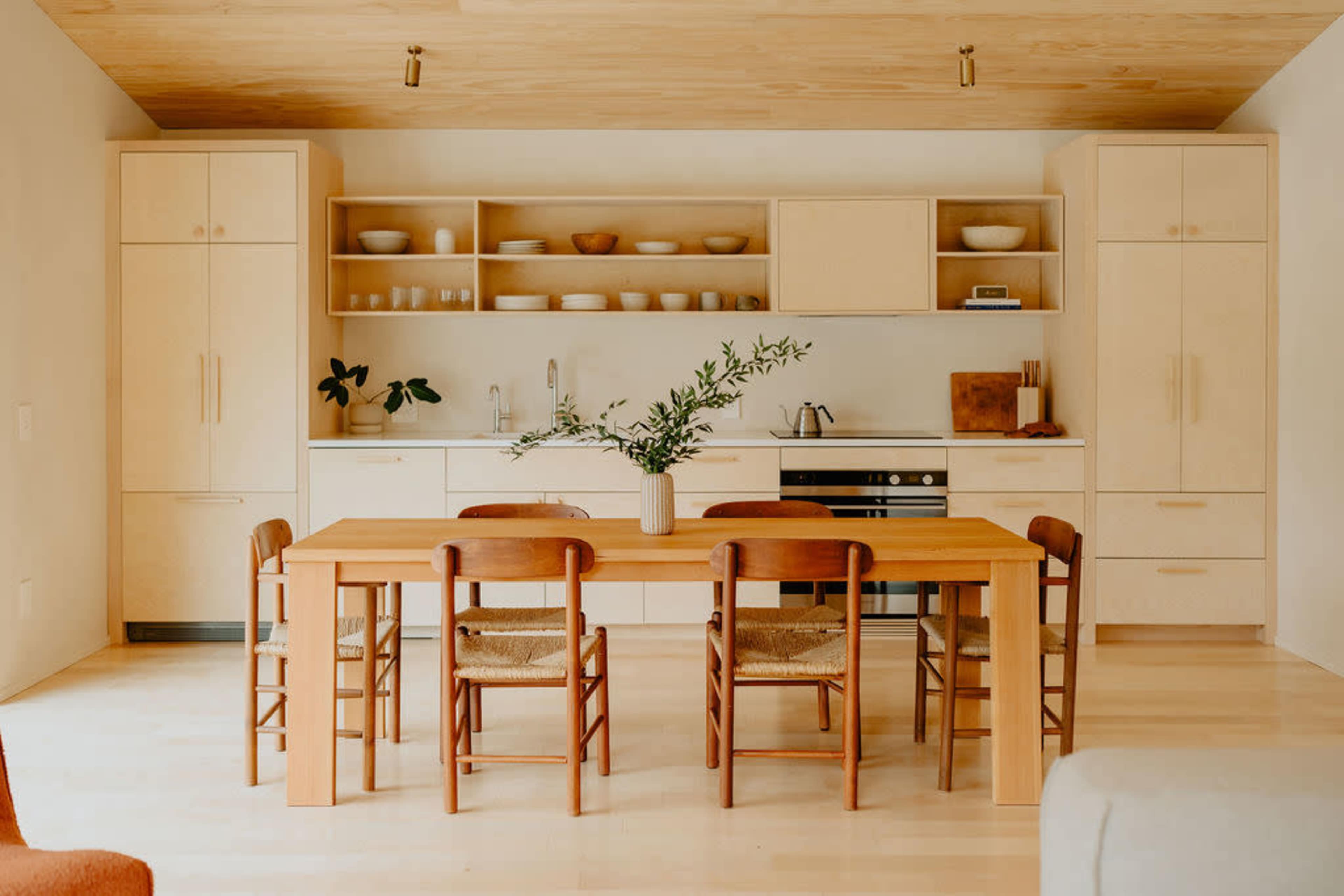 A modern kitchen and dining area features a wooden table surrounded by chairs, with light cabinetry, open shelves displaying dishware, and a kitchen counter.