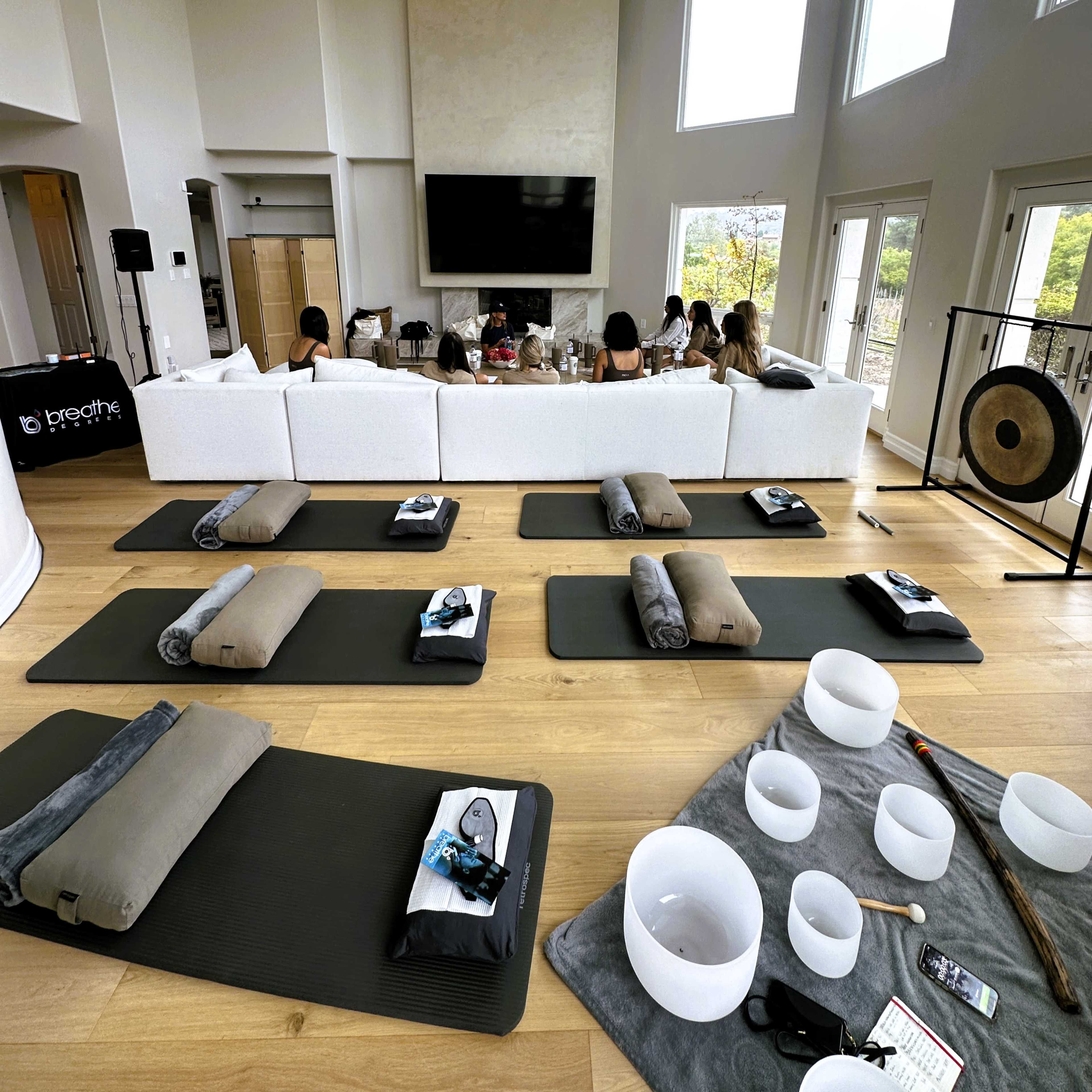 A spacious, modern living room is arranged for a wellness session, with mats and sound bowls set up on the floor and participants seated on a large white sectional couch.