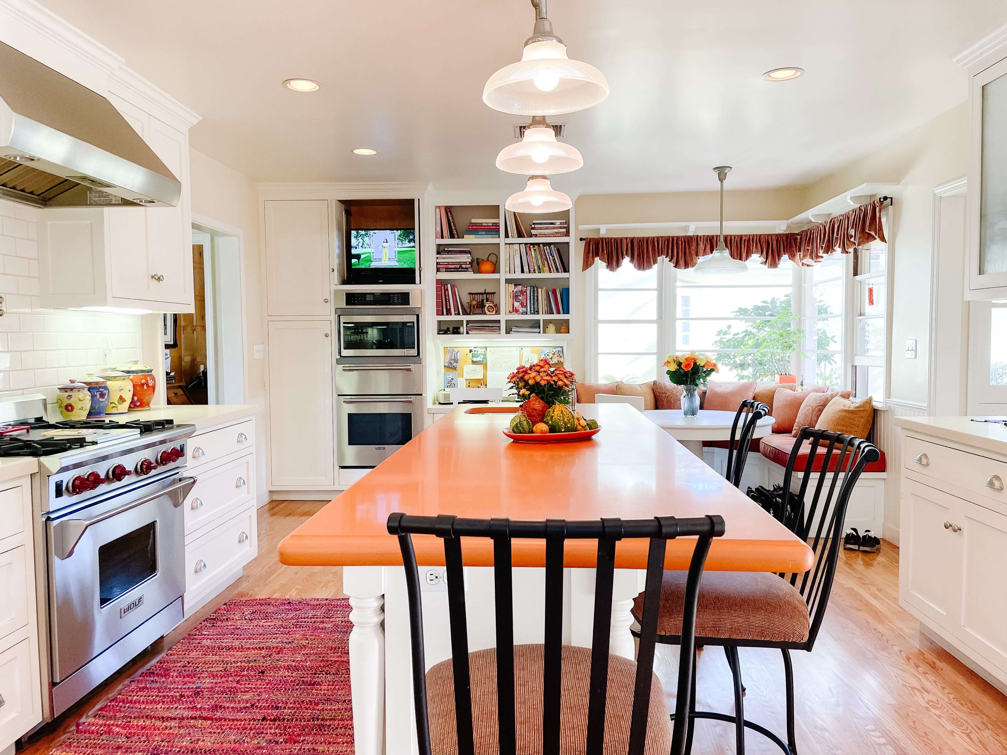 The kitchen features an orange countertop island surrounded by black bar stools, with white cabinetry and large windows providing natural light.