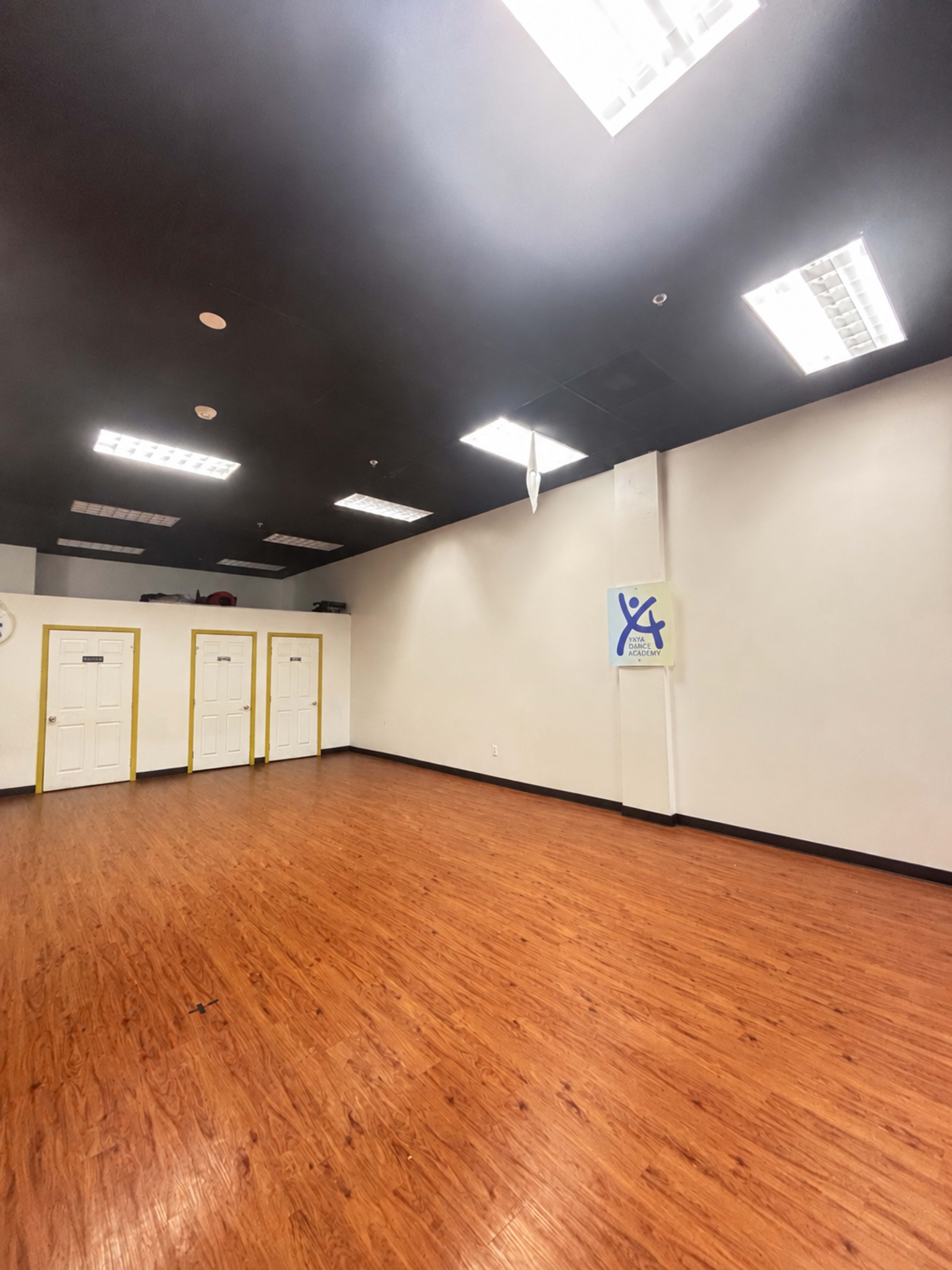 The image shows an empty room with wooden flooring, white walls, and three closed doors on one side.