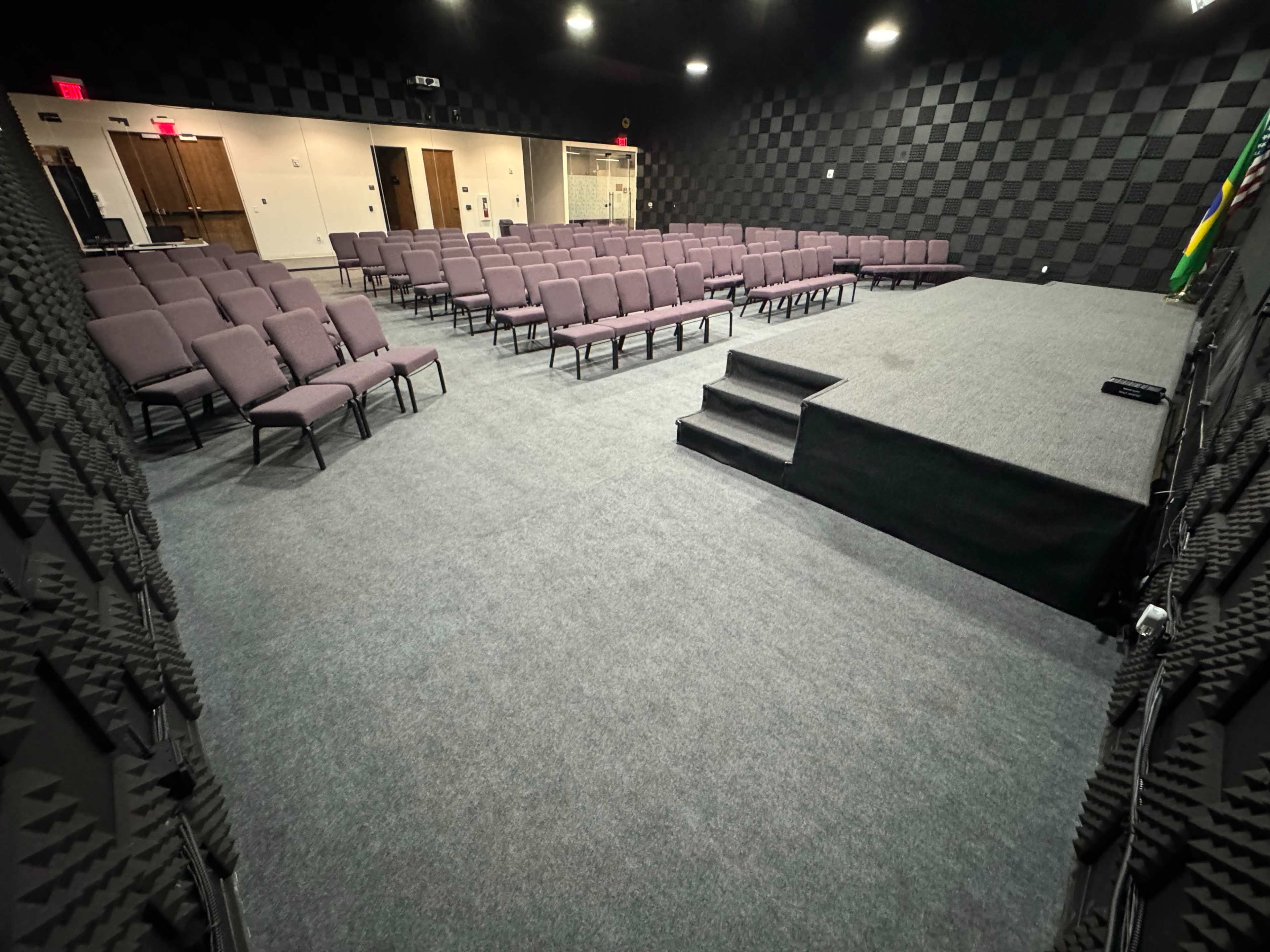 The image shows a conference room with rows of purple chairs arranged facing a small stage, all set on a gray carpeted floor.