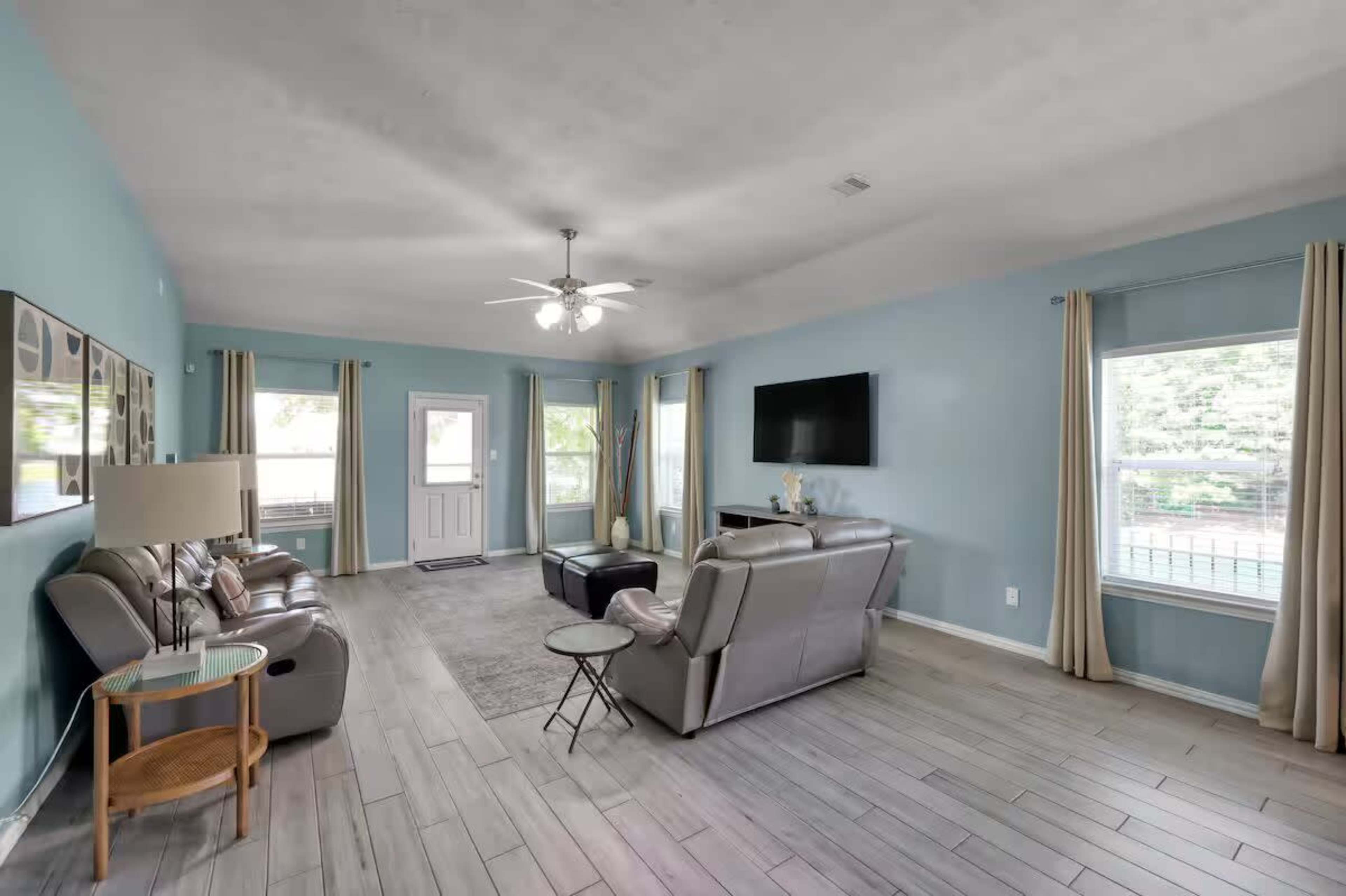The image depicts a spacious living room with light blue walls, two sofas, a coffee table, a ceiling fan, and large windows letting in natural light.