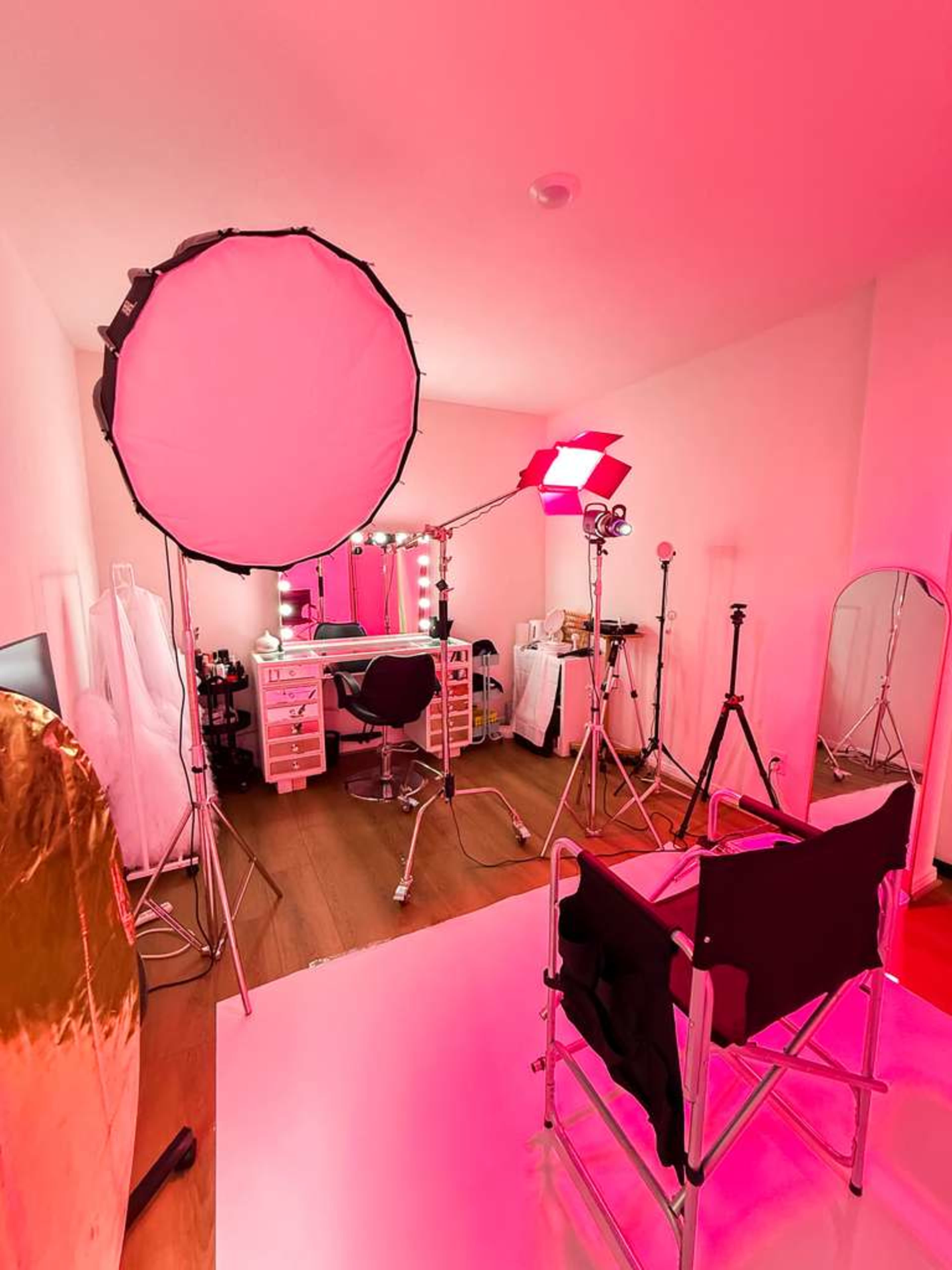The image shows a brightly lit makeup studio featuring a chair, lighting equipment, a mirror with vanity lights, and a large backdrop.