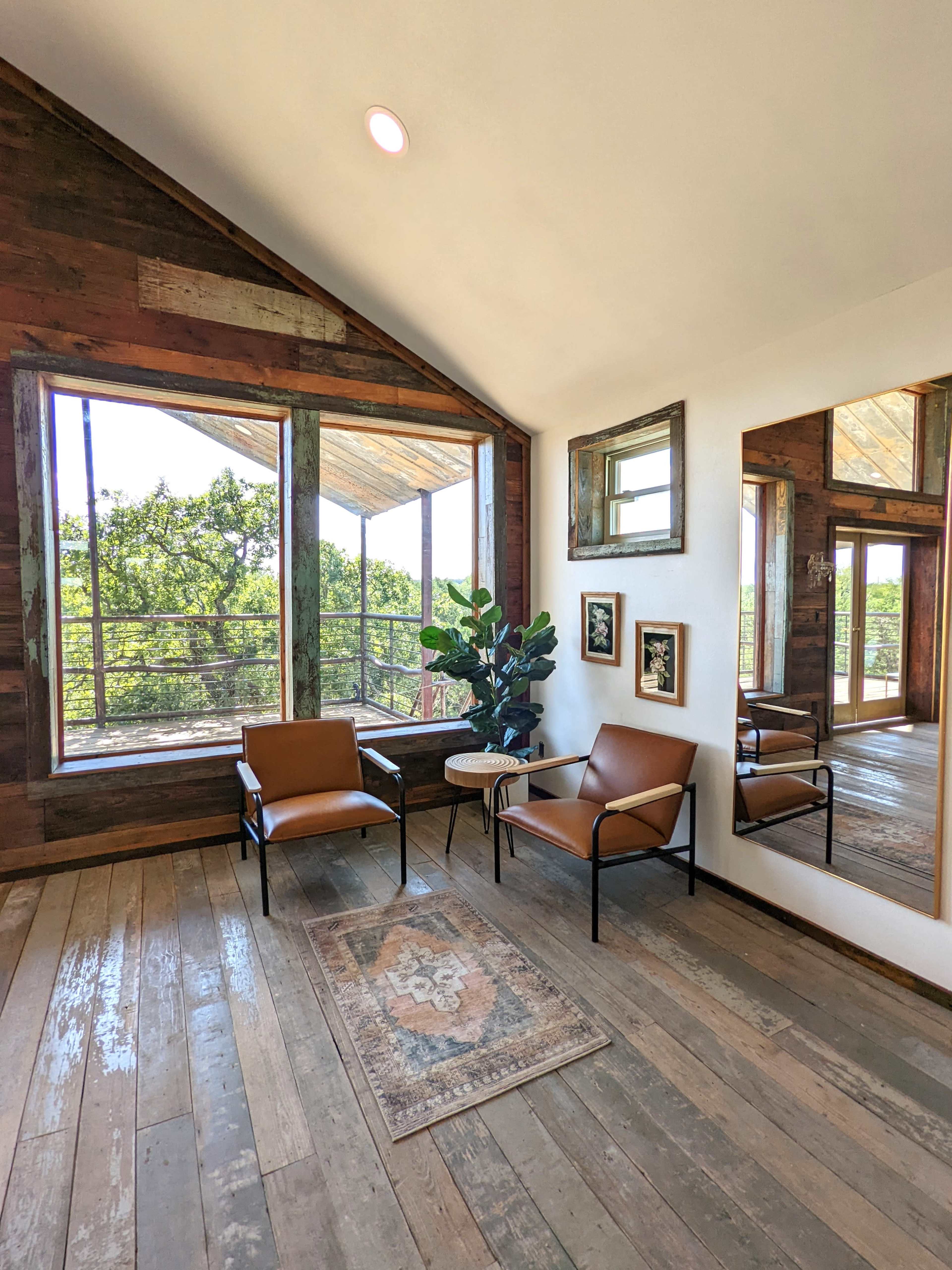 A living space with two brown chairs, a small side table, a potted plant, and large windows overlooking a scenic view.