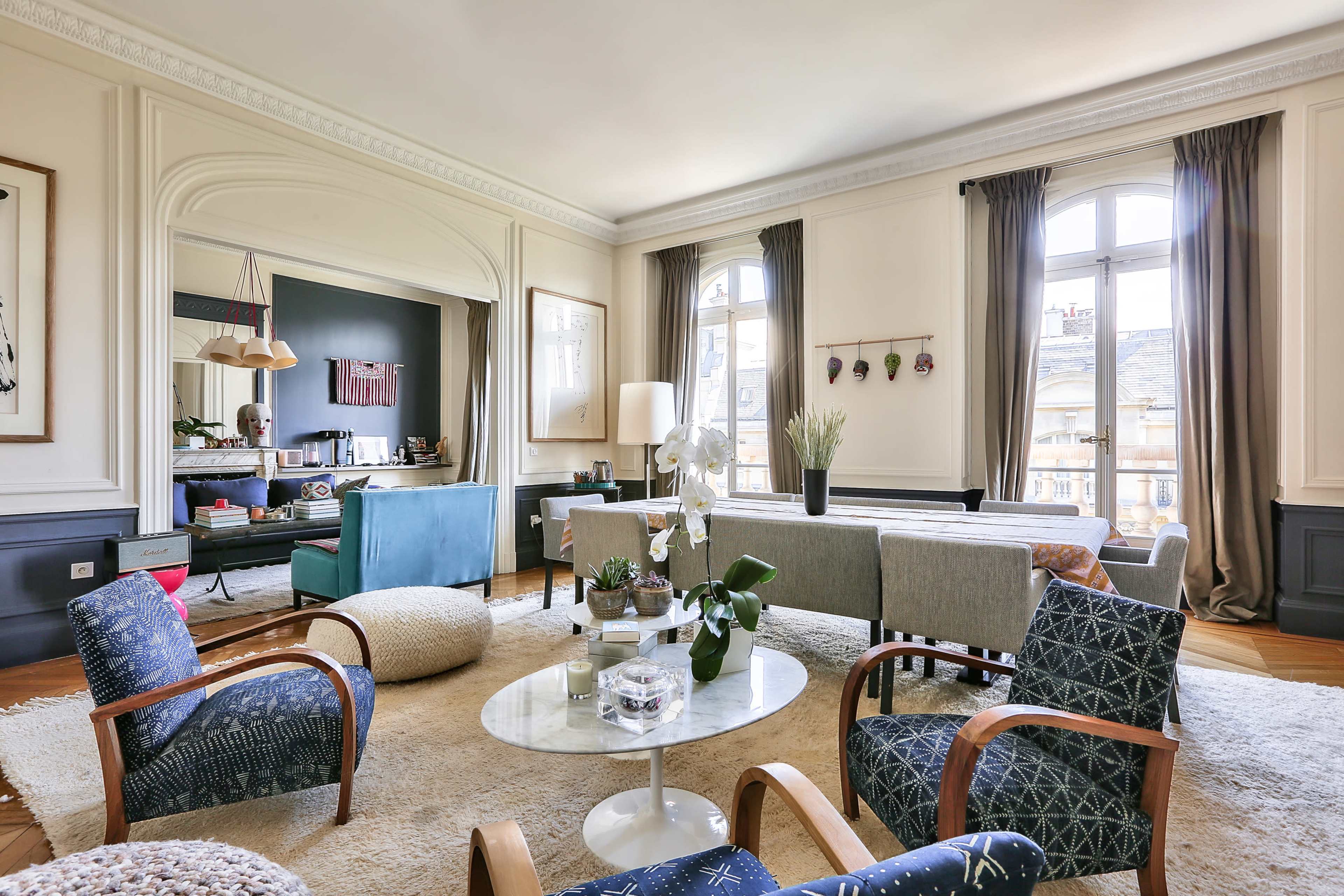 A bright and spacious living room featuring a mix of seating arrangements, a coffee table, and large windows with curtains.