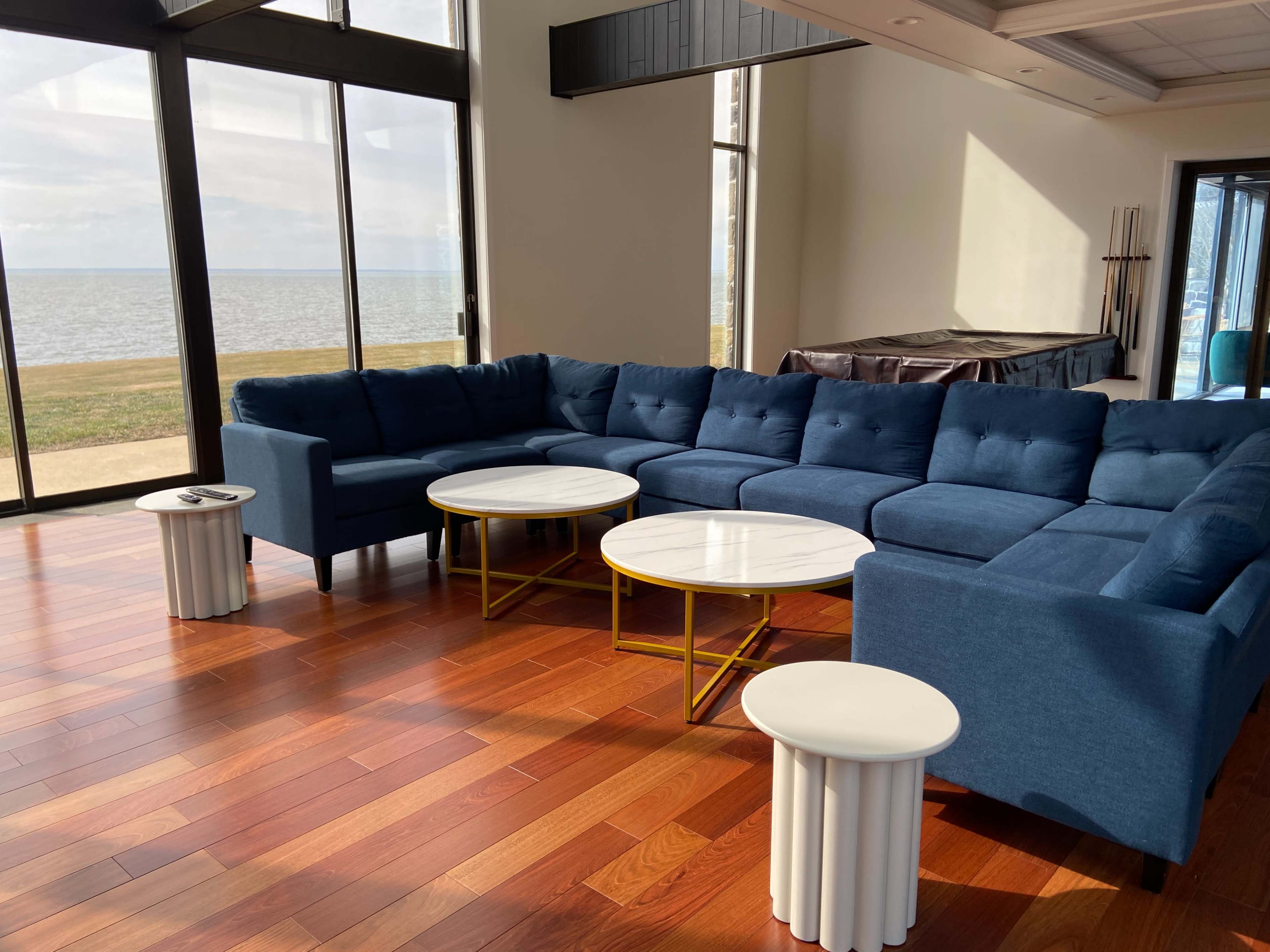 A large blue sectional sofa is arranged in a modern living room with two round coffee tables and large windows offering a view of a body of water.