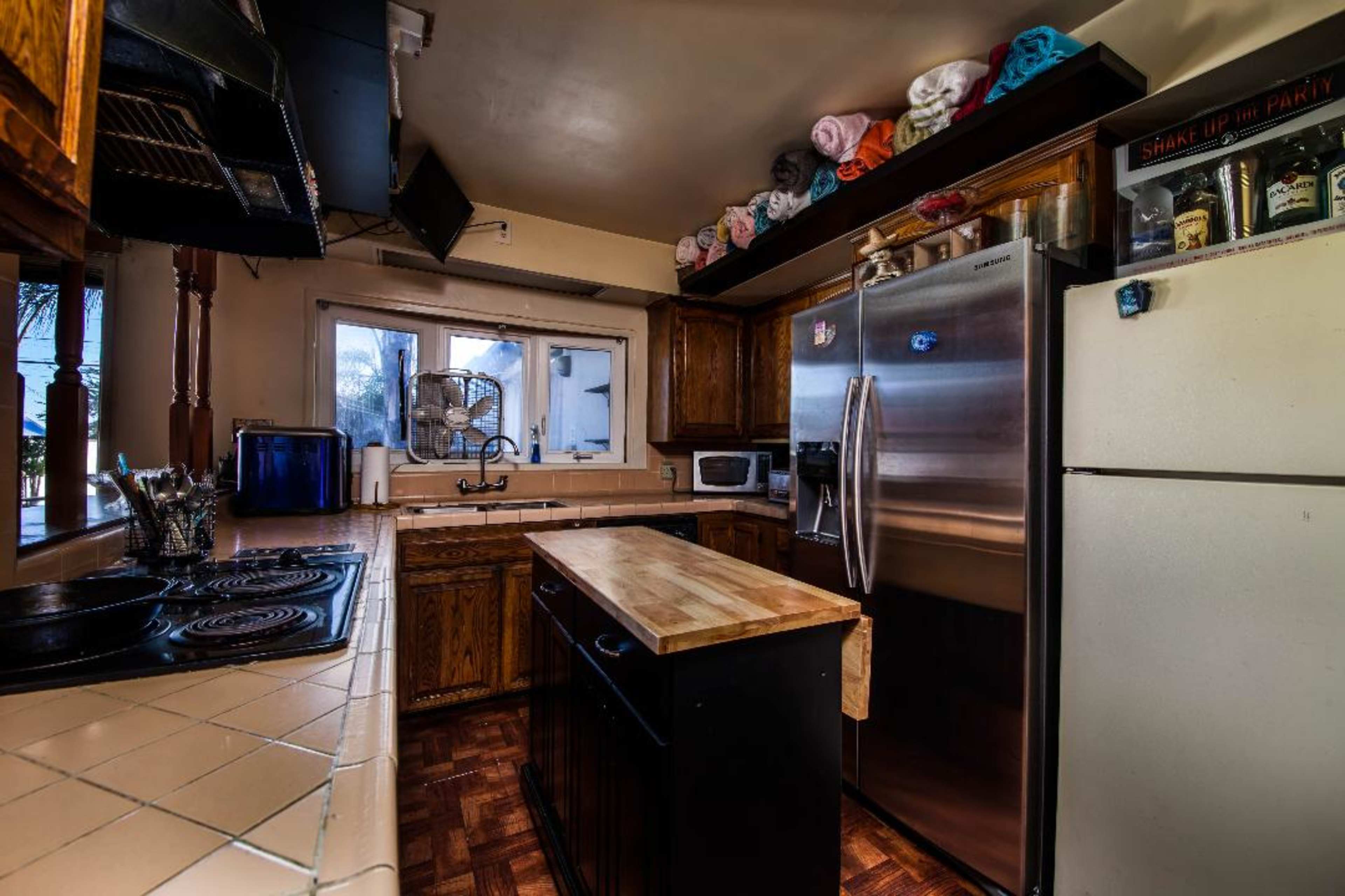 The kitchen features wooden cabinets, a central island with a butcher block top, and stainless steel appliances, with colorful towels arranged on shelves above.