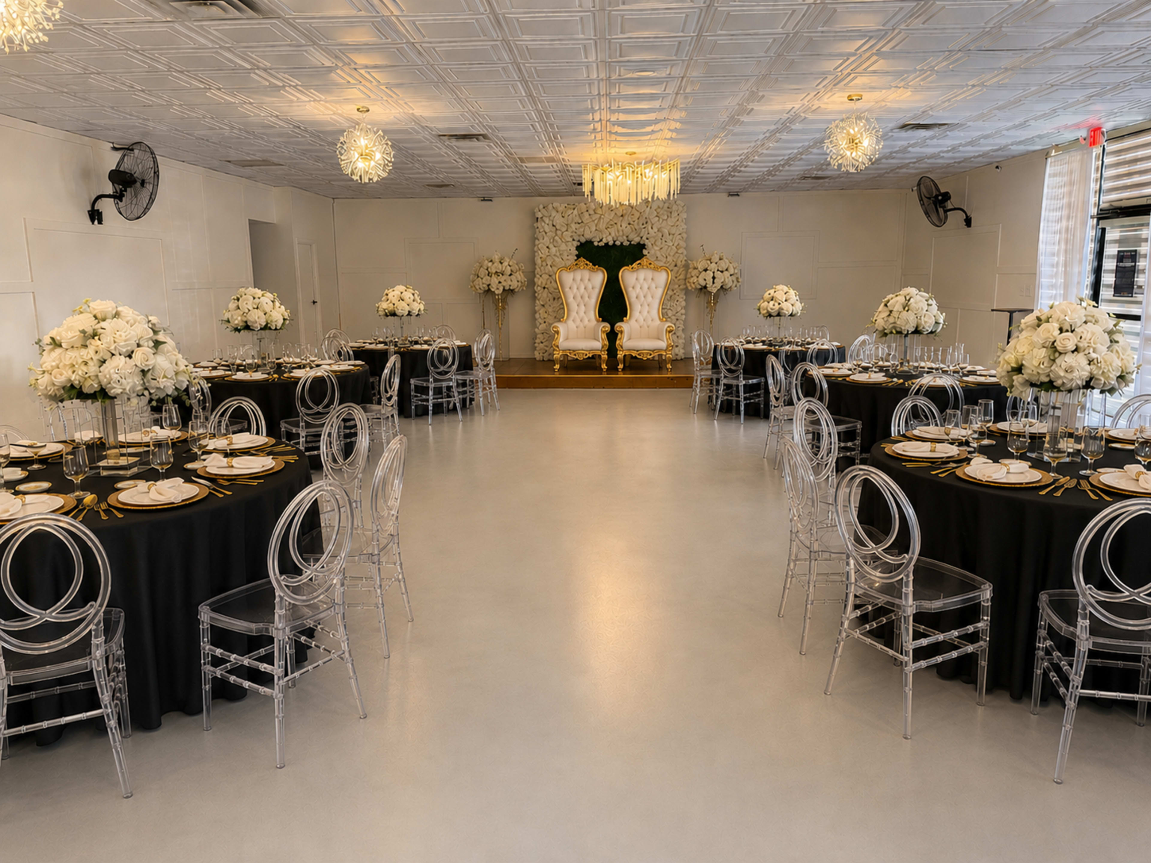 The image shows a decorated event space with circular tables set for dining, featuring clear acrylic chairs and floral centerpieces, along with a backdrop for a stage area.
