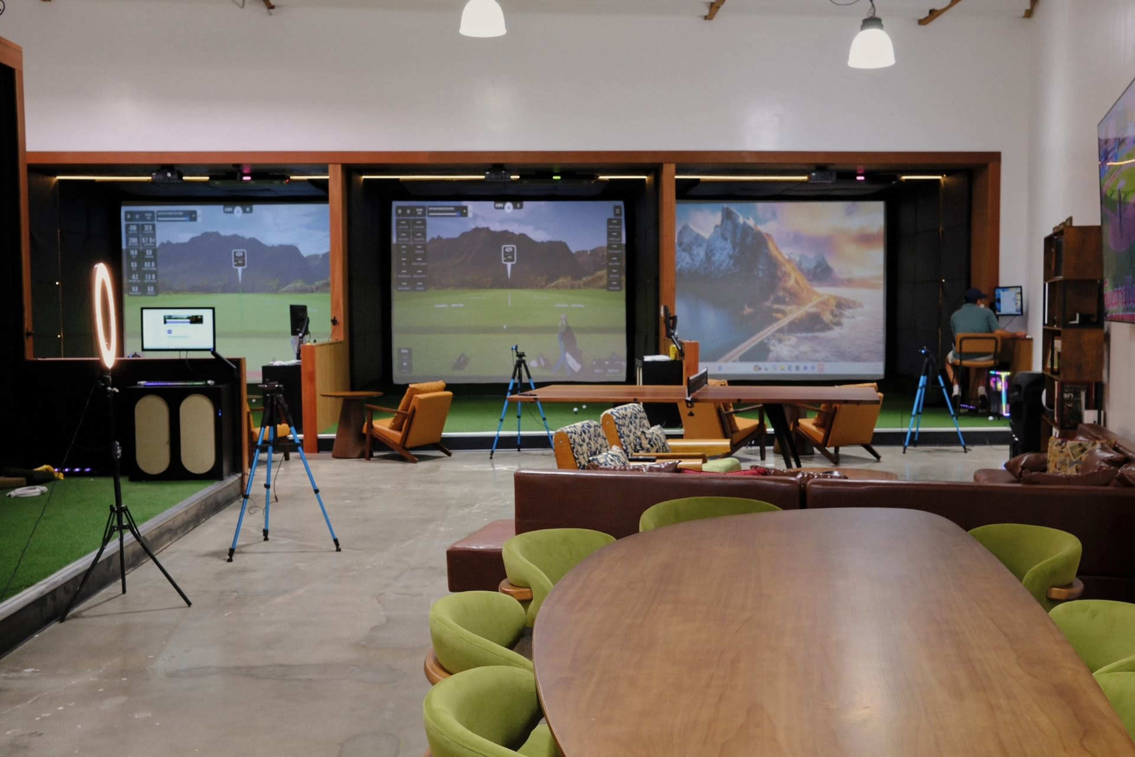 The image shows a spacious indoor setup for golf simulation, featuring multiple projection screens and various equipment positioned around a large central table and seating area.