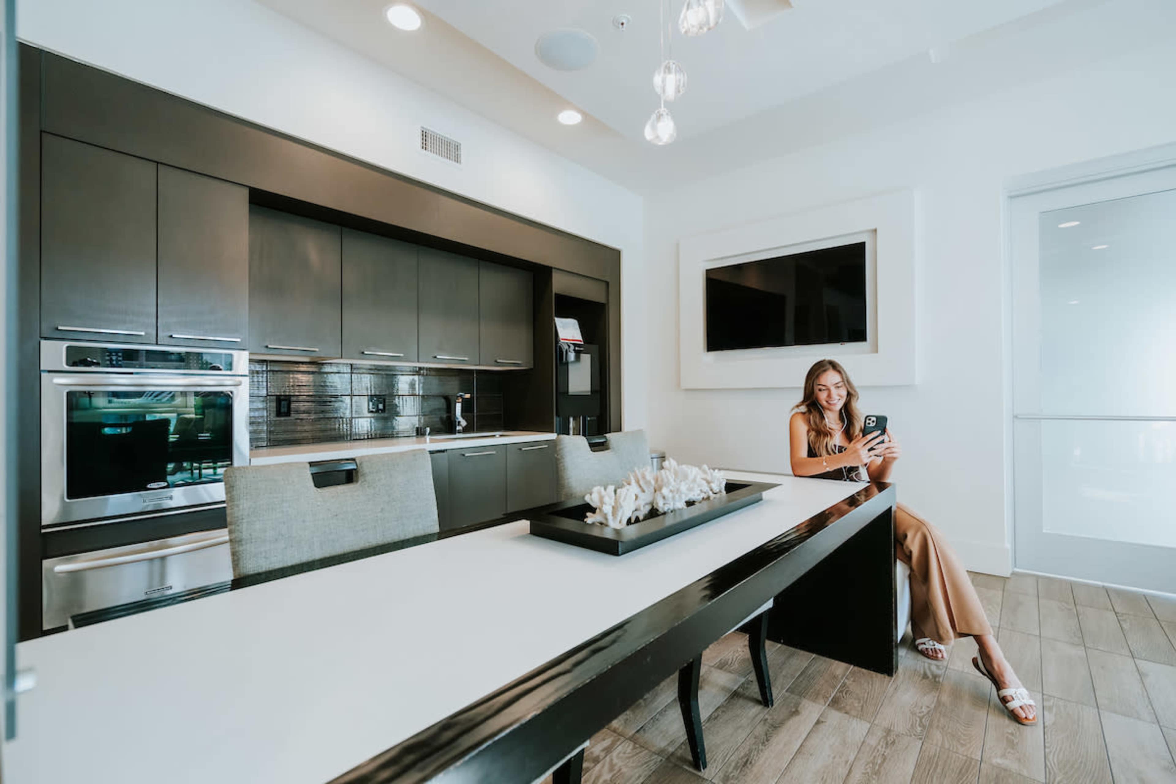 A woman sits at a long dining table in a modern kitchen with dark cabinetry and a large wall-mounted television.