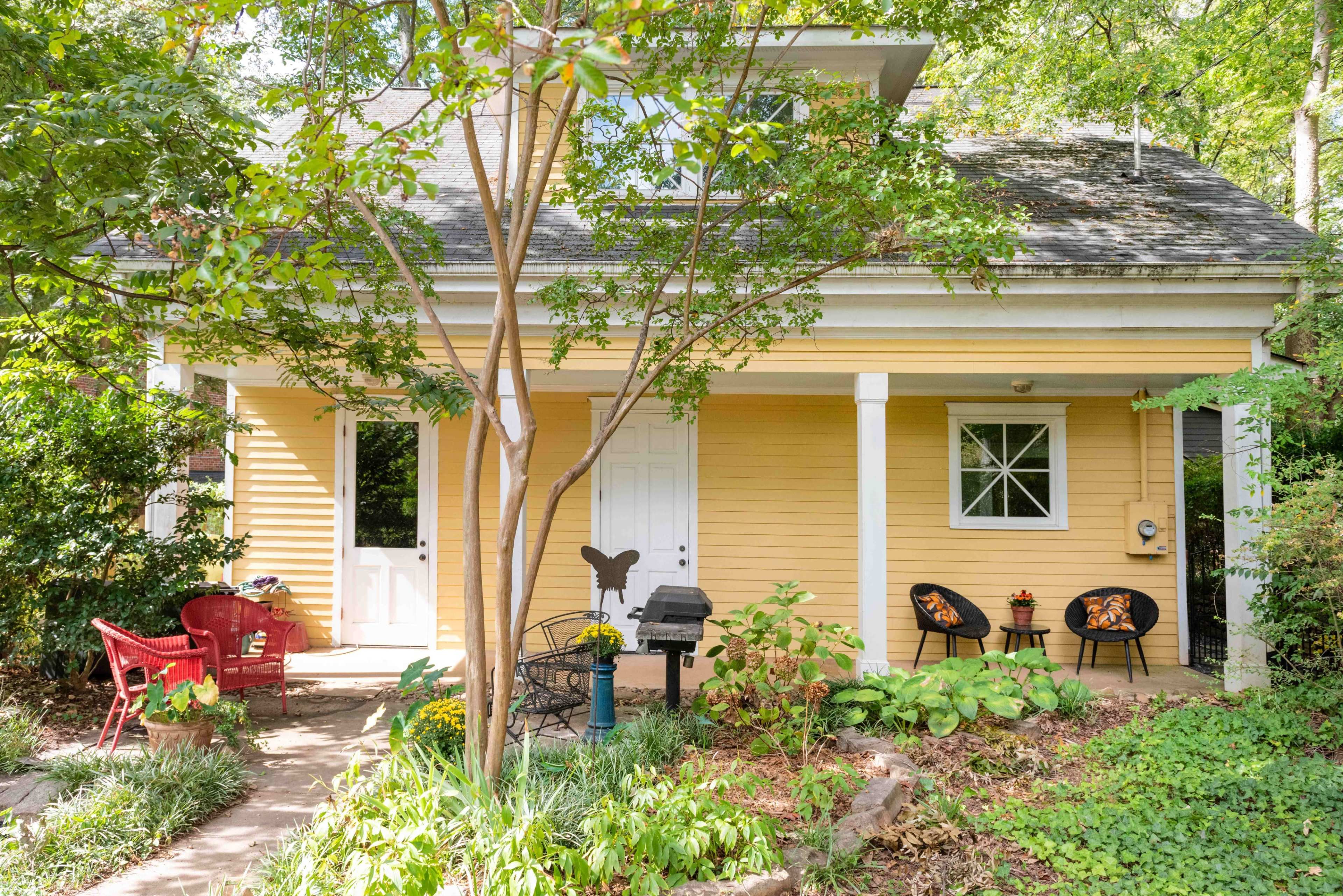 A yellow house with a porch features seating areas and a grill, surrounded by lush greenery.