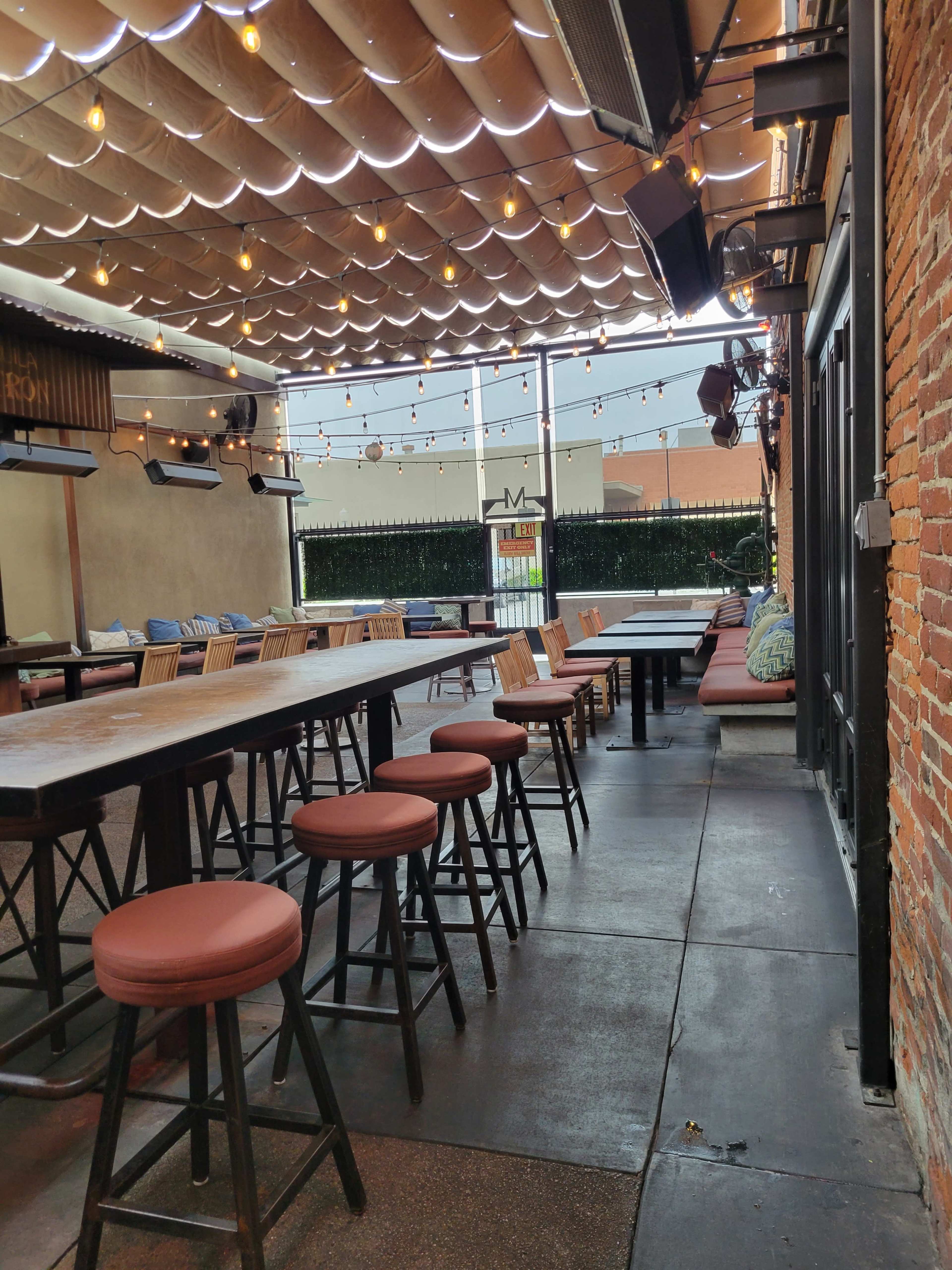 An indoor-outdoor restaurant with wooden tables, bar stools, and string lights overhead, featuring brick walls and a partially open roof.