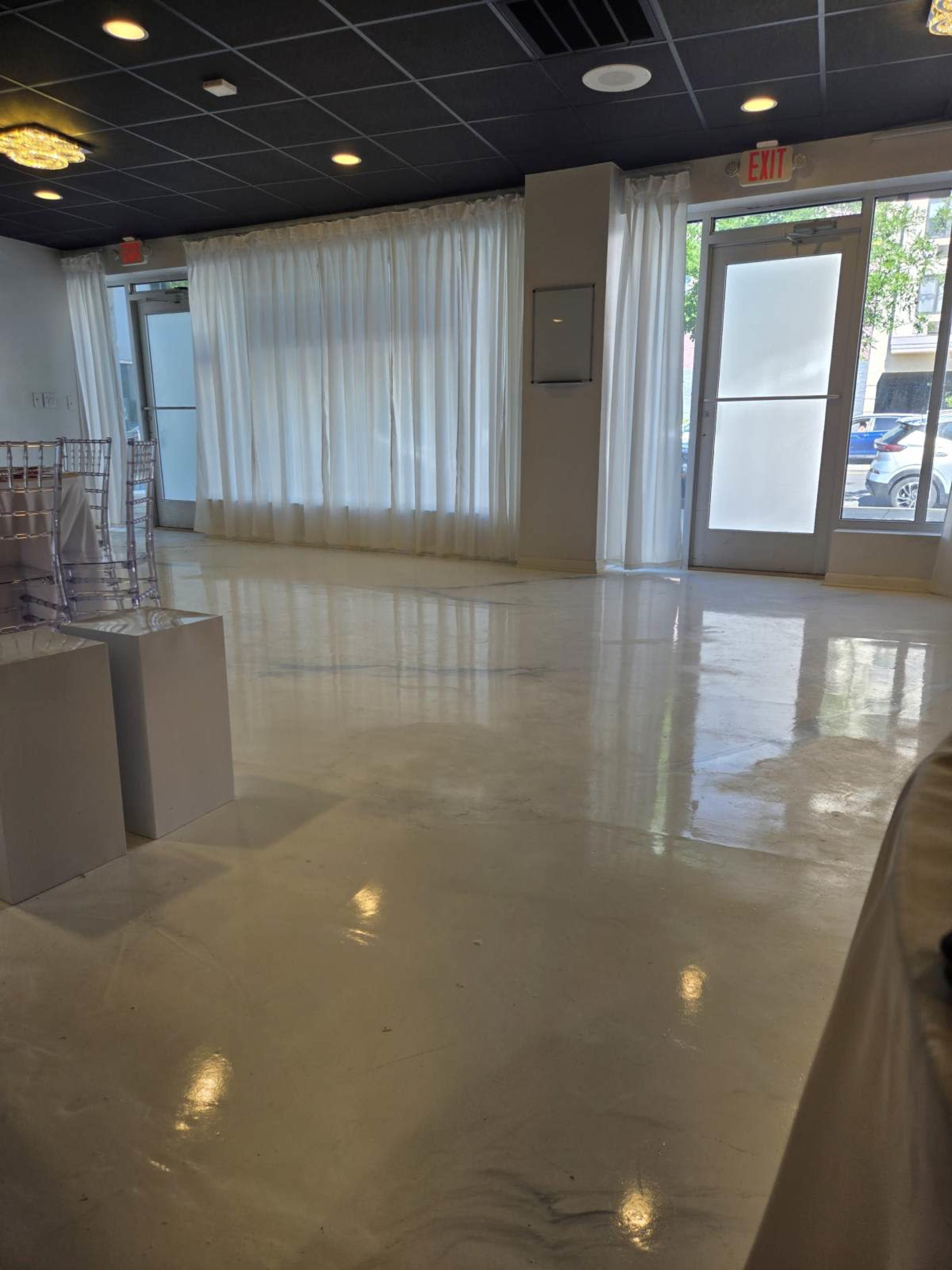 The image shows a spacious indoor area with white walls, polished flooring, large windows covered by sheer curtains, and double glass doors leading outside.