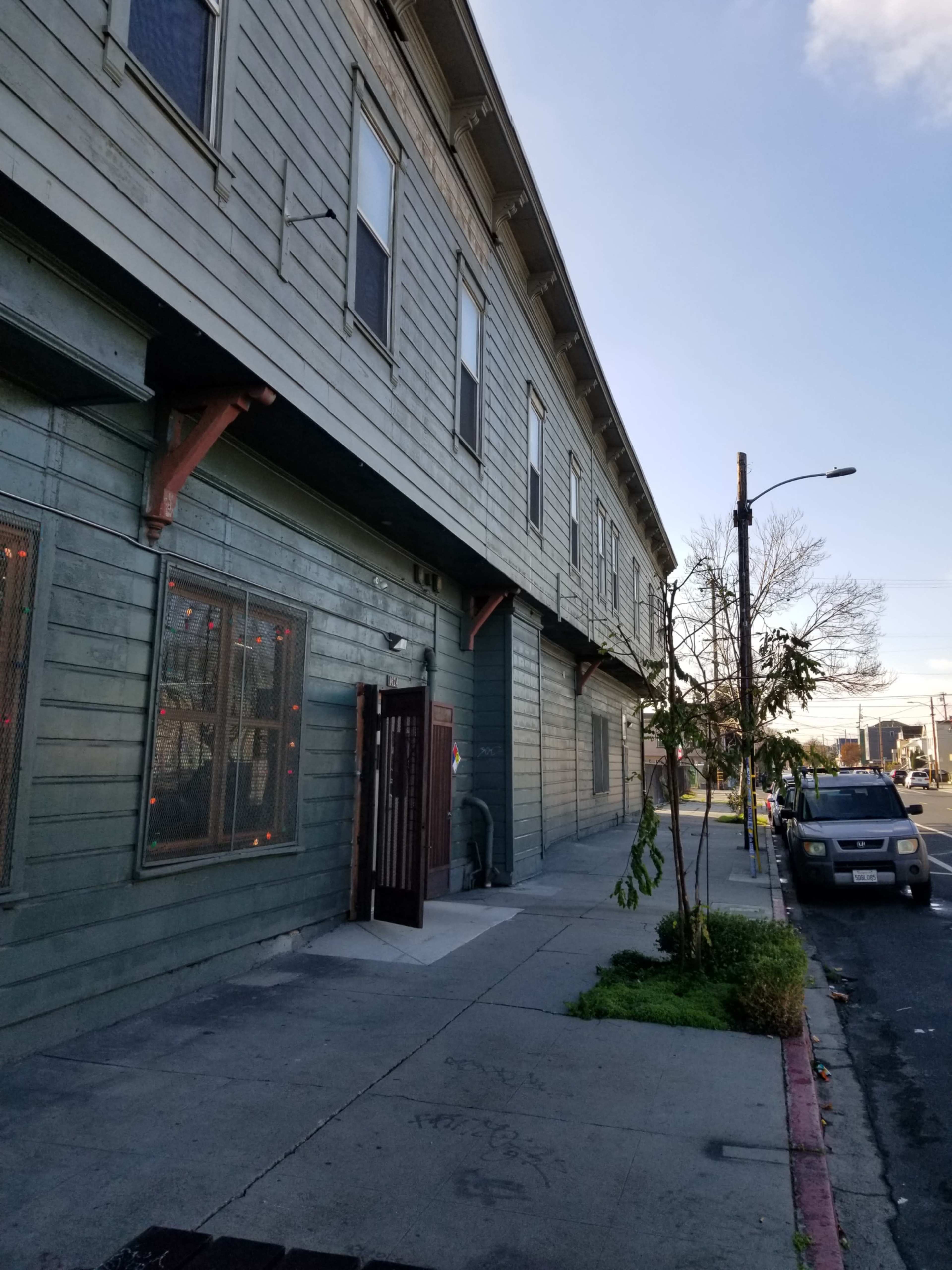 The image shows the side of a green wooden building with visible windows and a door, alongside a sidewalk that features a small tree and a parked car.