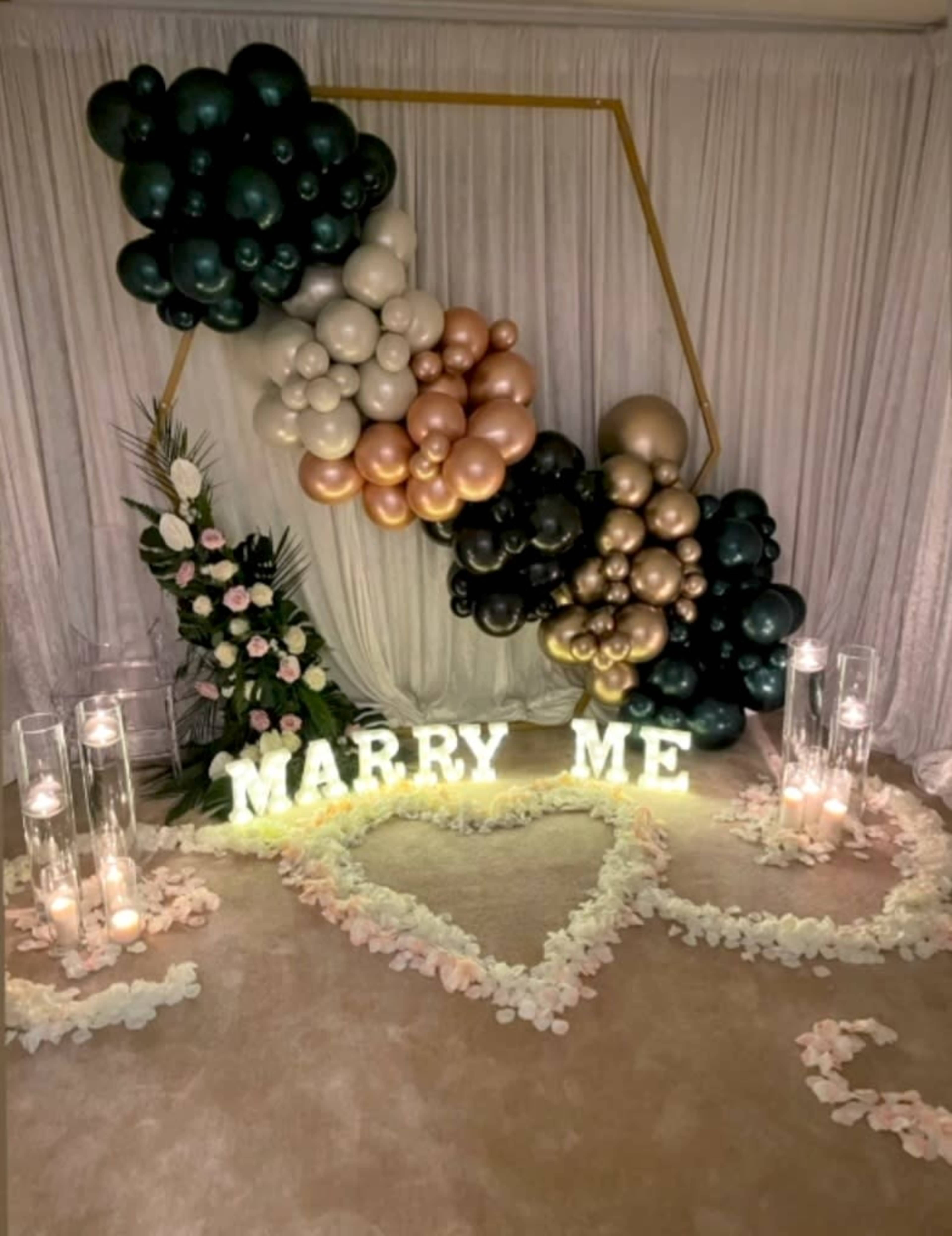 A decorative setup for a proposal, featuring a heart-shaped arrangement of flower petals, illuminated letters spelling "MARRY ME," and a backdrop of balloons in various colors.