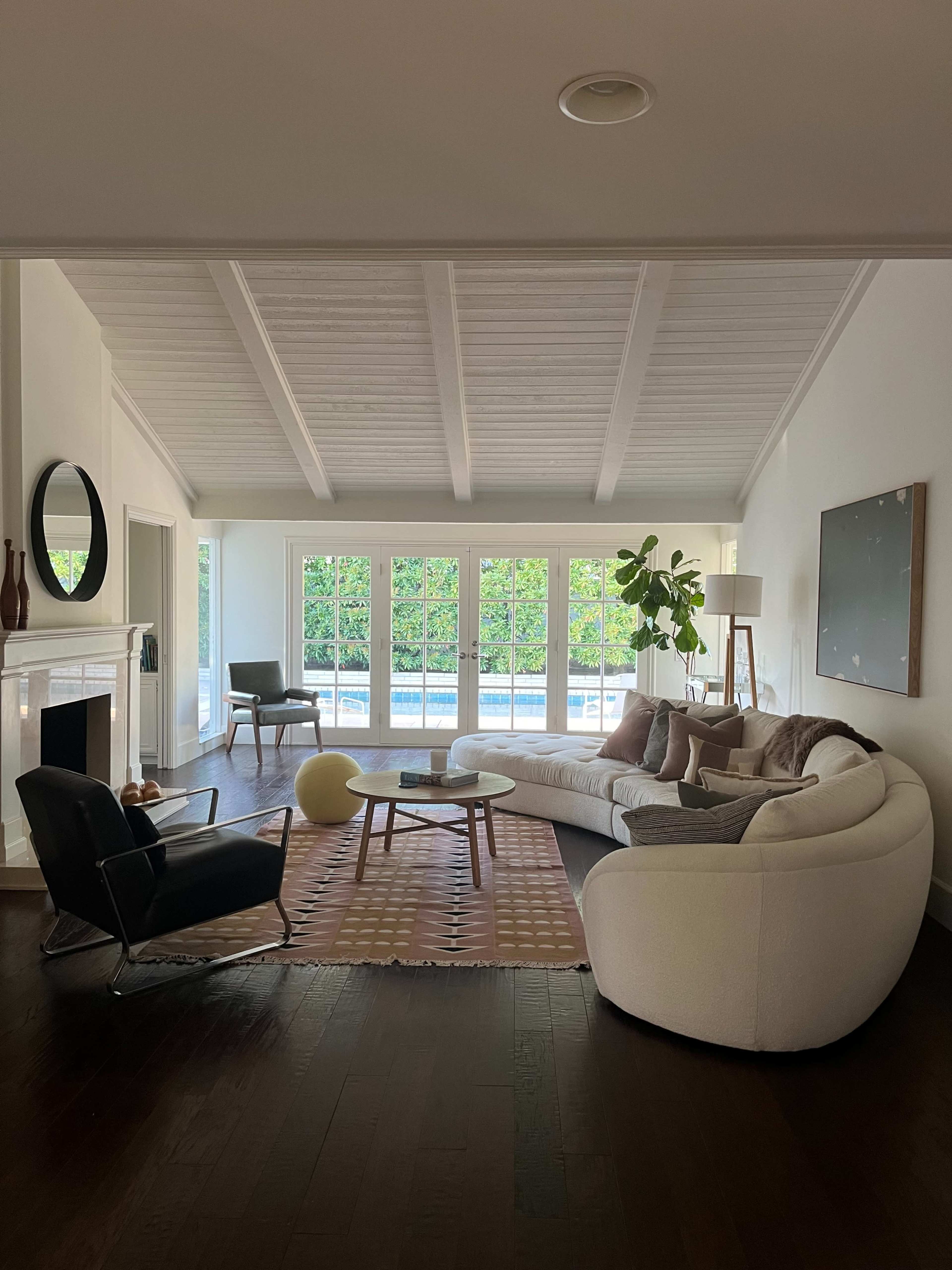 A modern living room features a curved white sofa, a black armchair, and large windows overlooking a pool, with a cozy rug and decorative plants.
