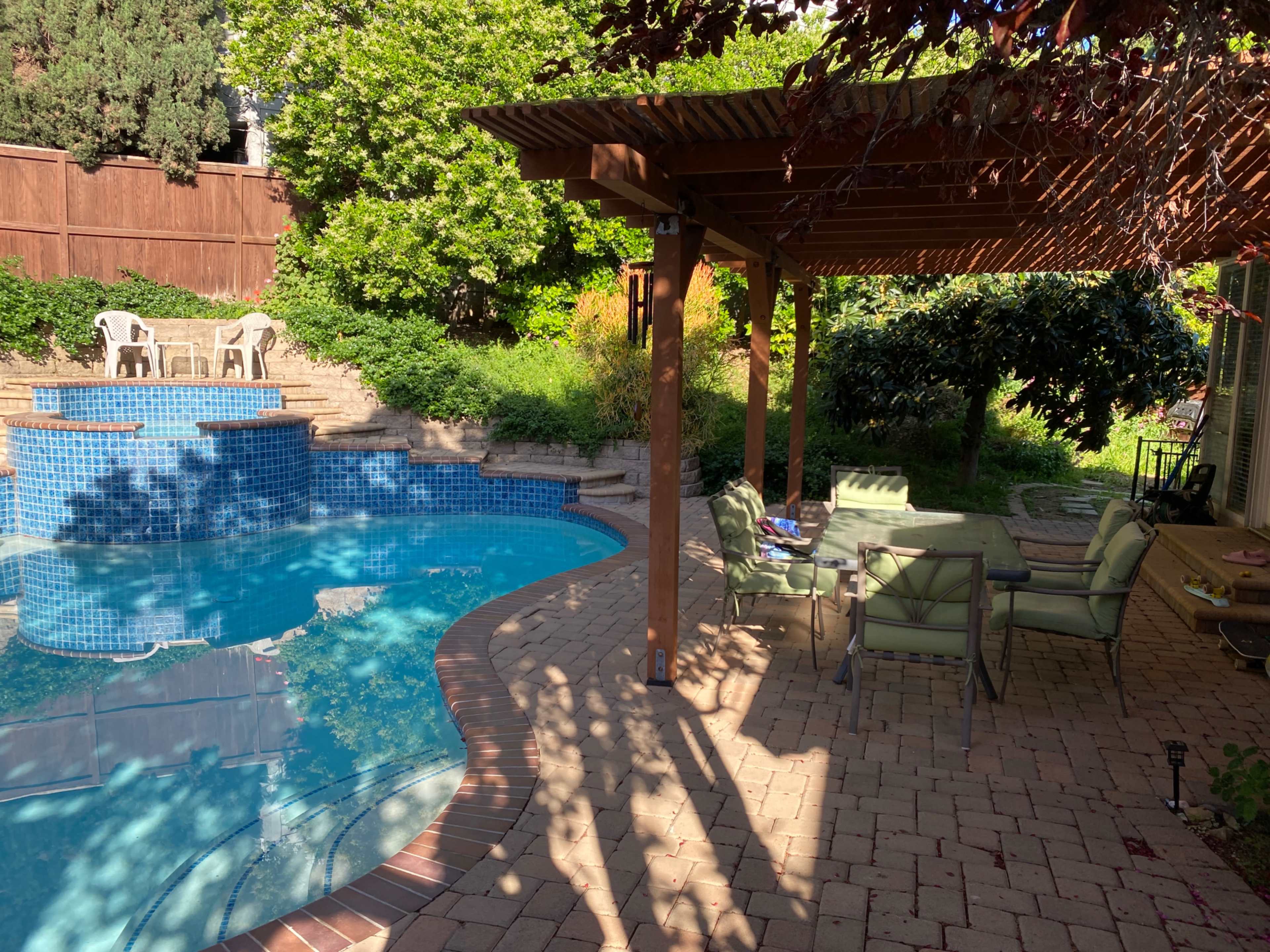 The image shows a backyard scene featuring a swimming pool surrounded by greenery, a wooden pergola, and outdoor seating.