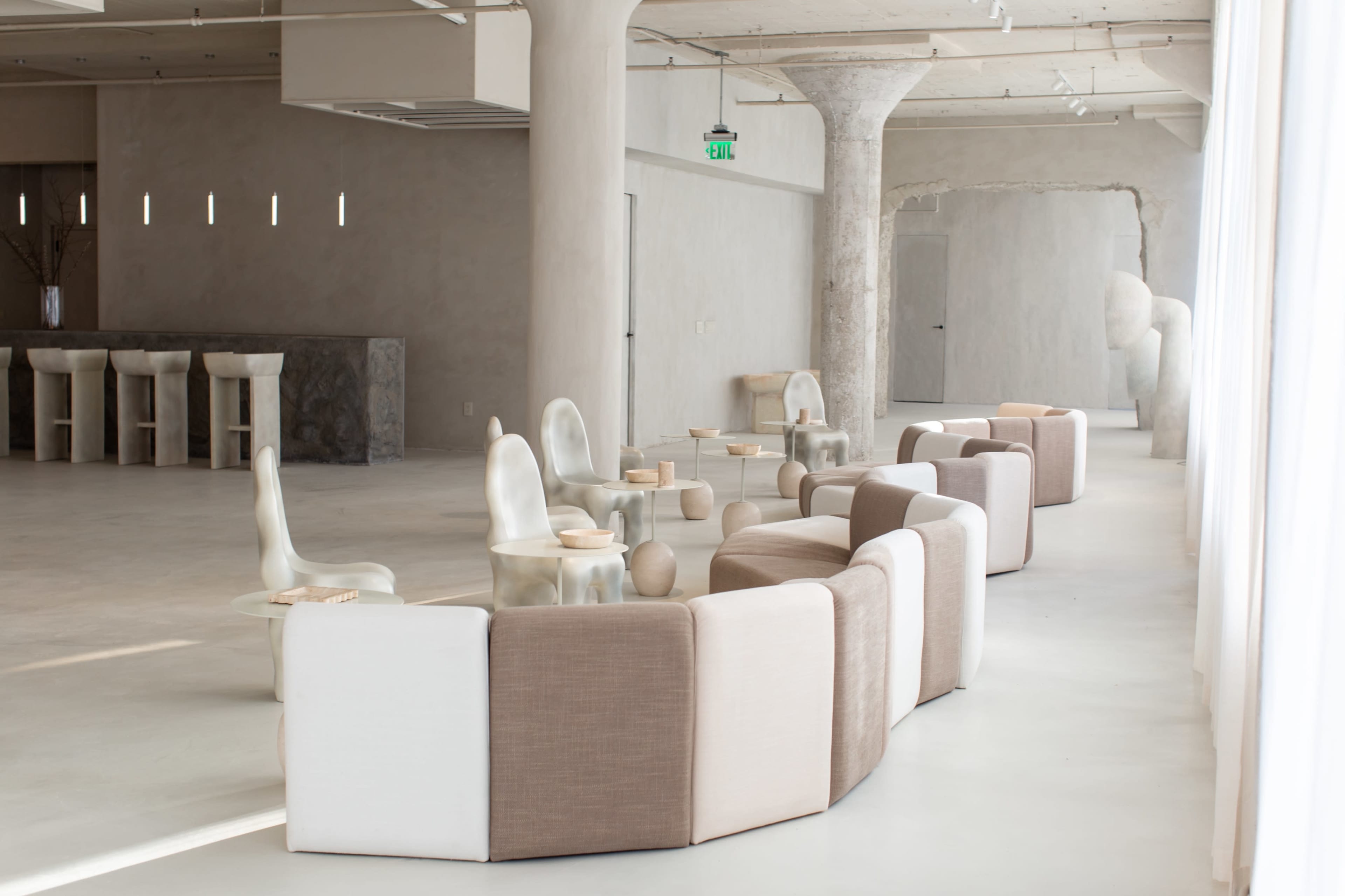 The image shows a modern interior space featuring a curved sofa arrangement and several tables, with minimalist decor and large windows allowing natural light.