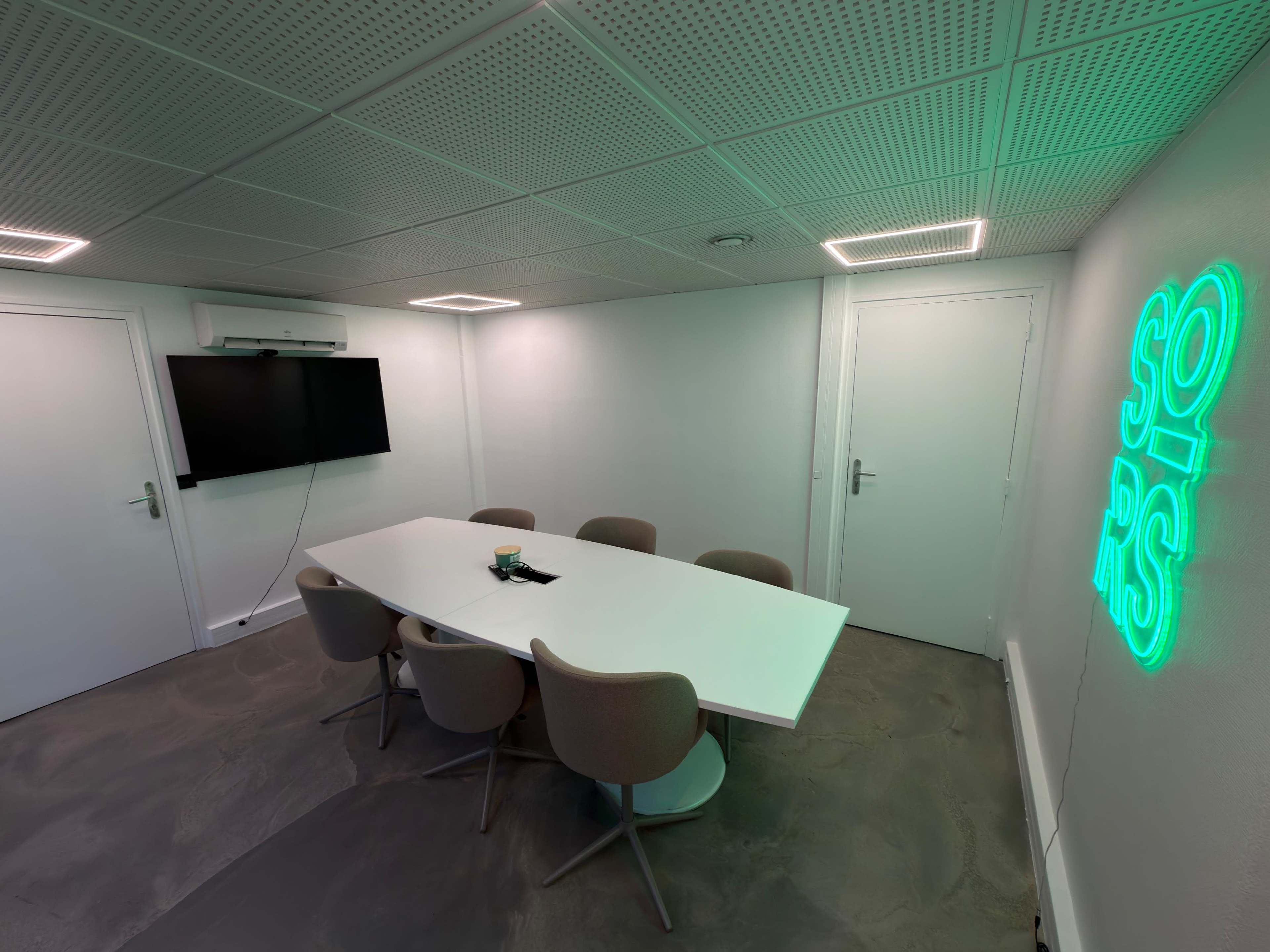 A modern conference room features a white table surrounded by beige chairs, with a wall-mounted TV and illuminated green neon signage.