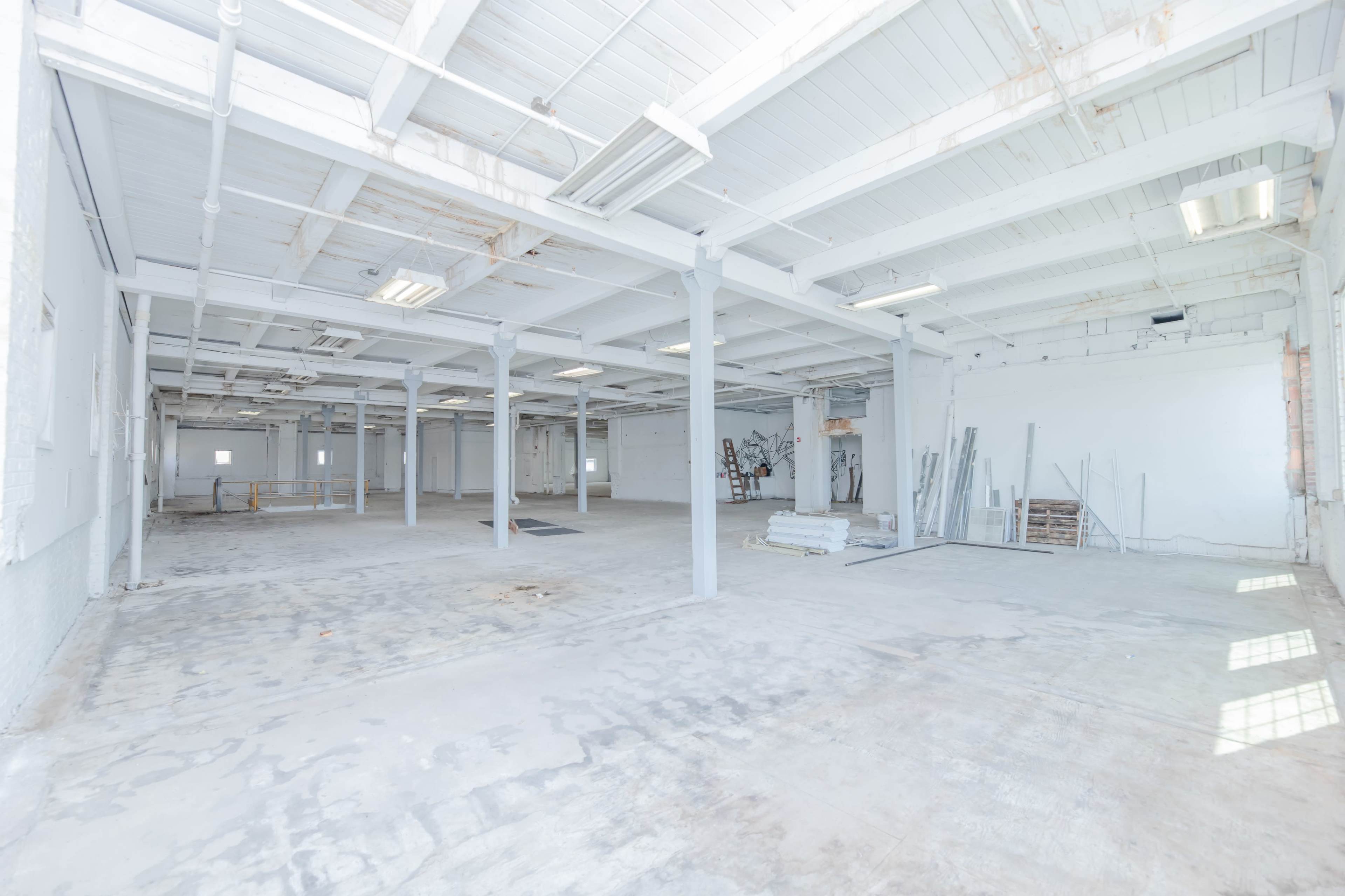 A large, empty industrial space with concrete floors and white walls, featuring exposed beams and minimal furnishings.
