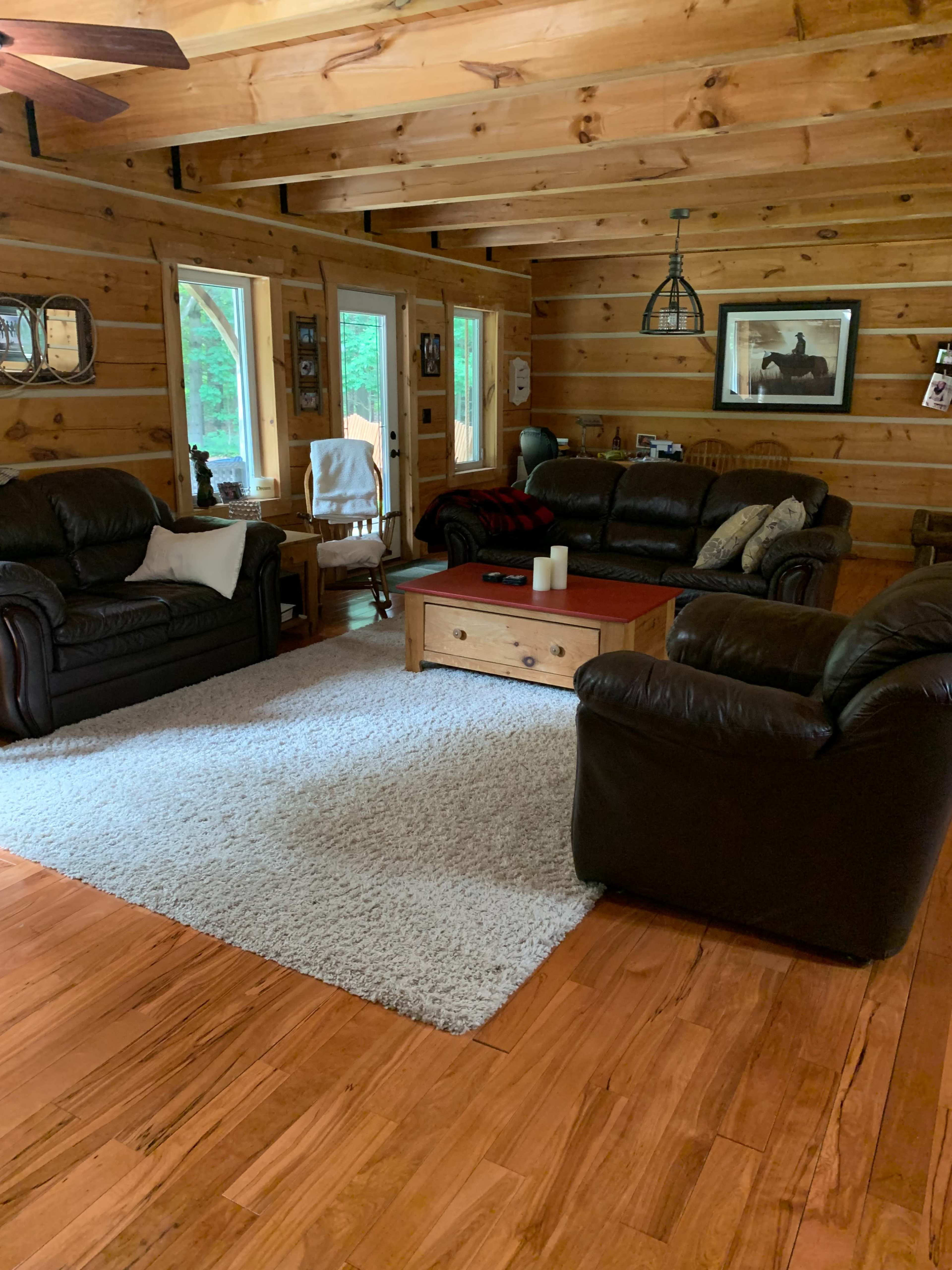 The room features wooden walls and flooring, with two leather sofas, a coffee table, and a large area rug, illuminated by natural light from large windows.