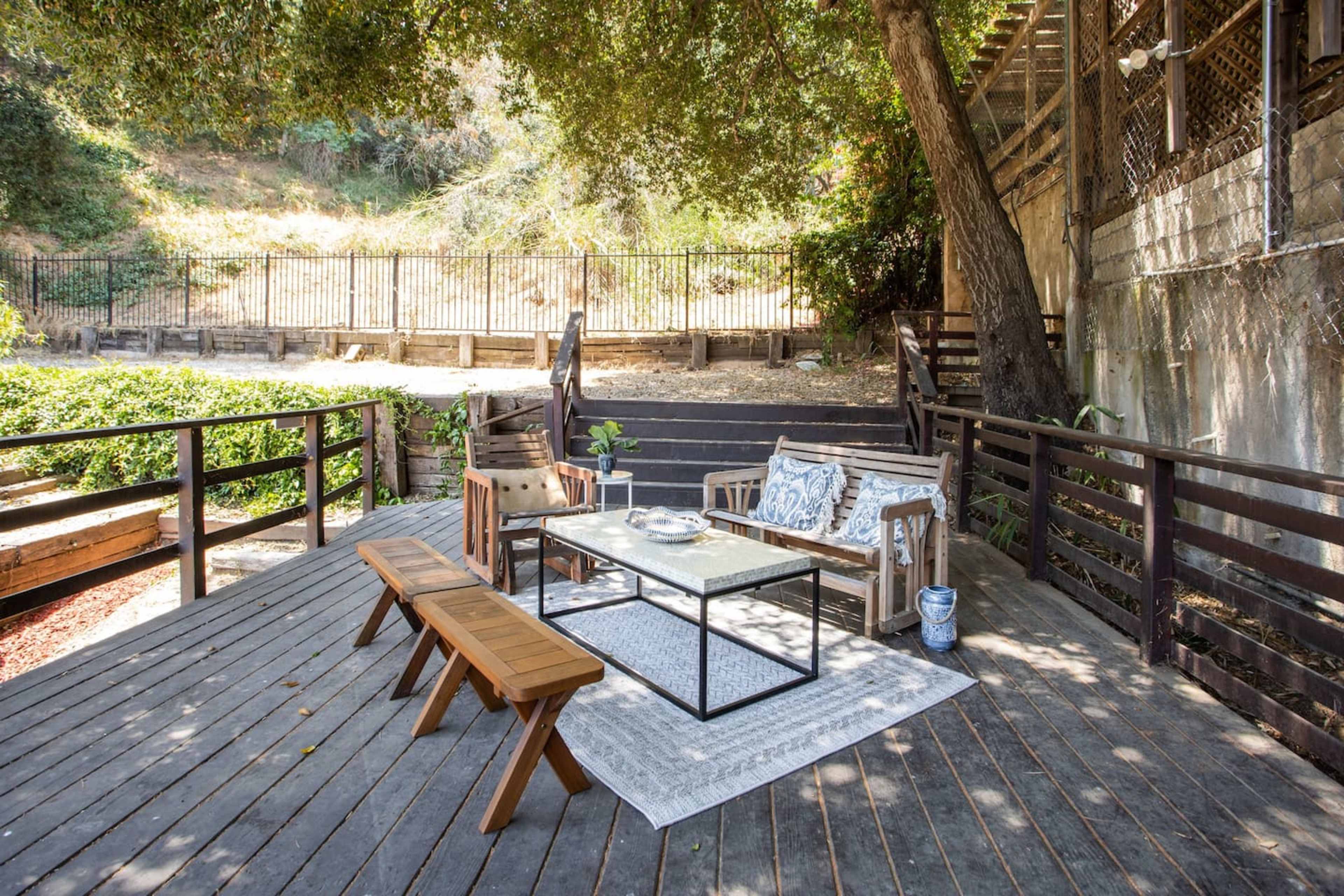 The image shows a wooden deck area furnished with a couch, chairs, a coffee table, and a bench, surrounded by trees and a fence.