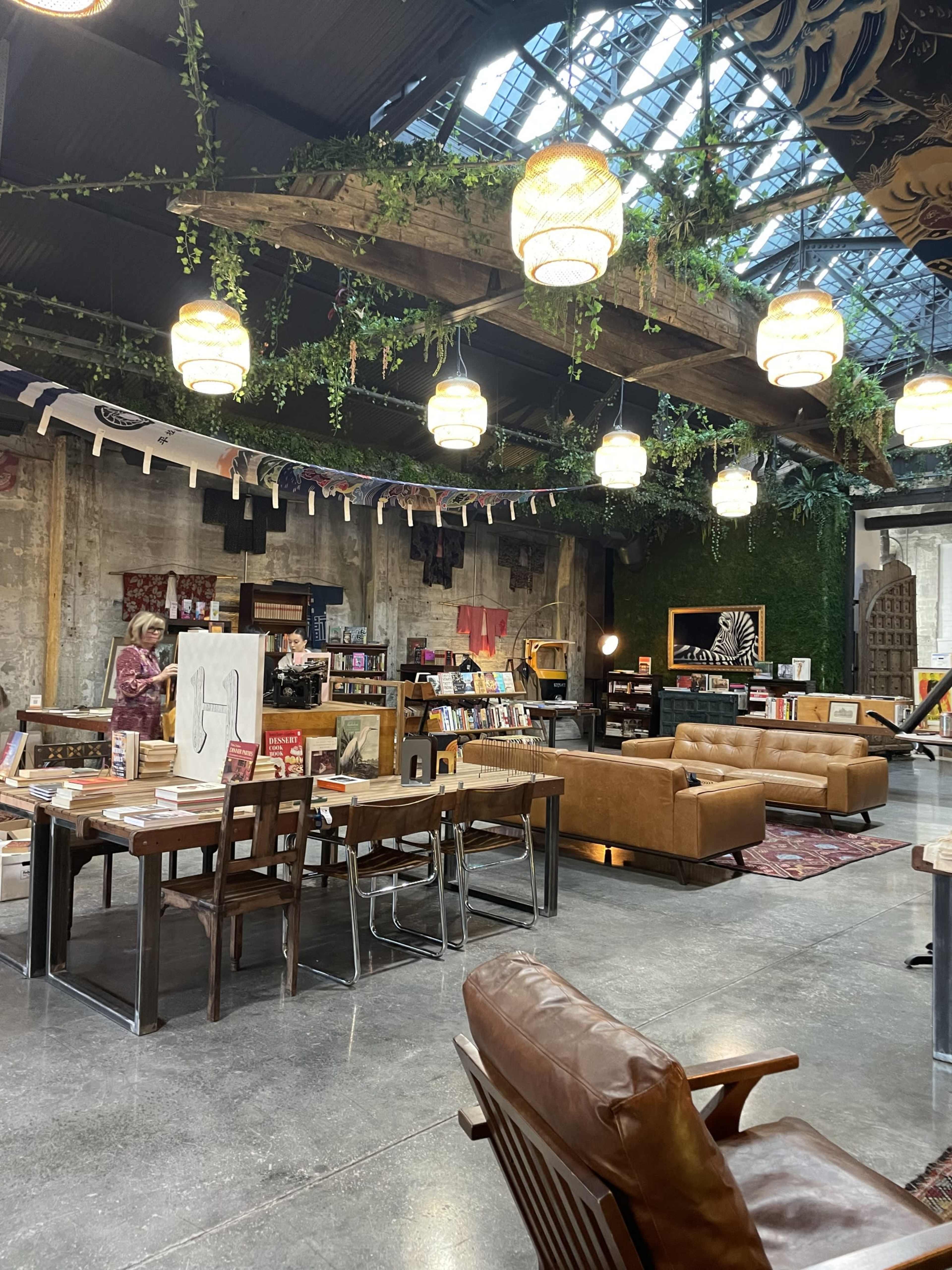 The image shows a stylish indoor space with seating areas, wooden tables, bookshelves, and decorative lighting, featuring a mix of greenery and artistic displays.