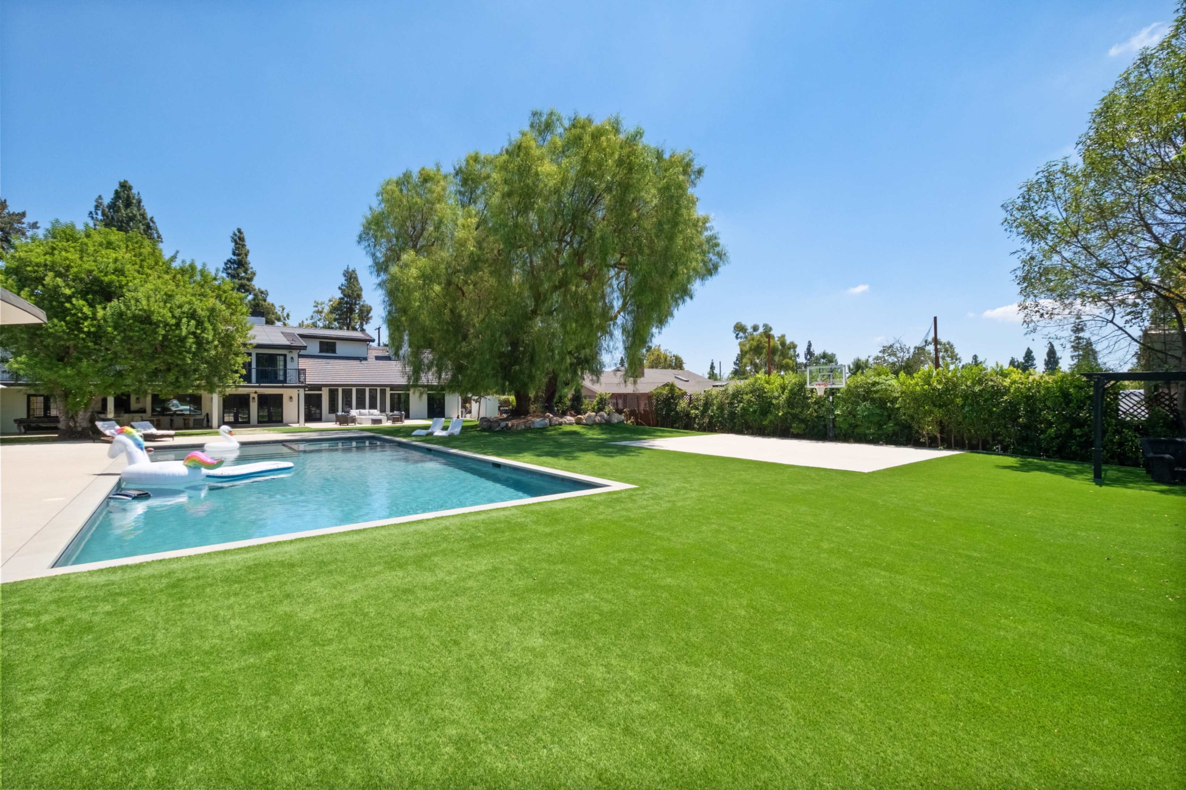 The image shows a spacious backyard featuring a swimming pool, a well-manicured lawn, and trees surrounding a modern home.