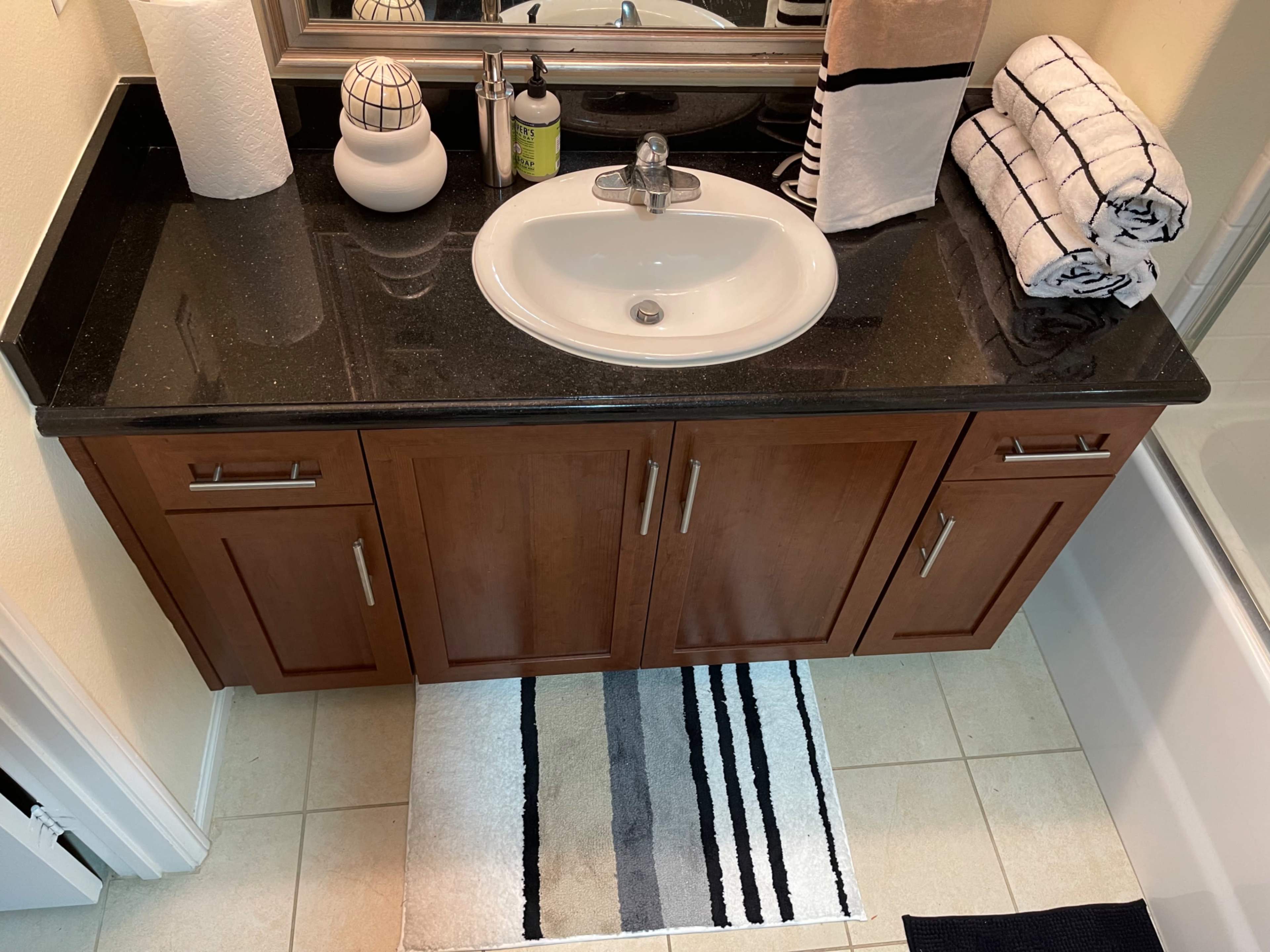 A bathroom vanity with a sink, dark countertop, and wooden cabinets is shown, accompanied by rolled towels and a striped rug.