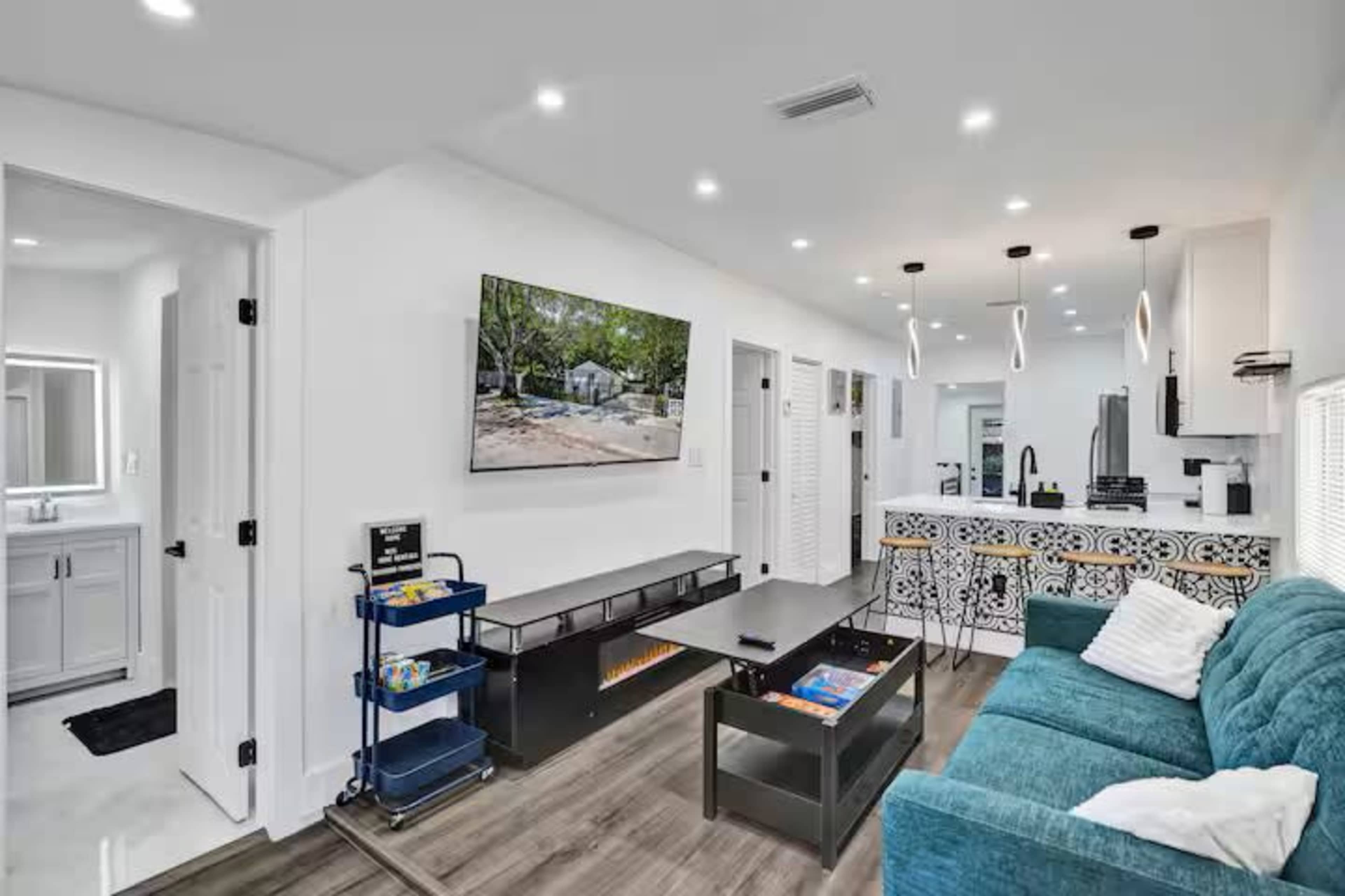 The image shows a modern living room with a teal couch, a wall-mounted television, and a kitchen area featuring a decorative tile backsplash.