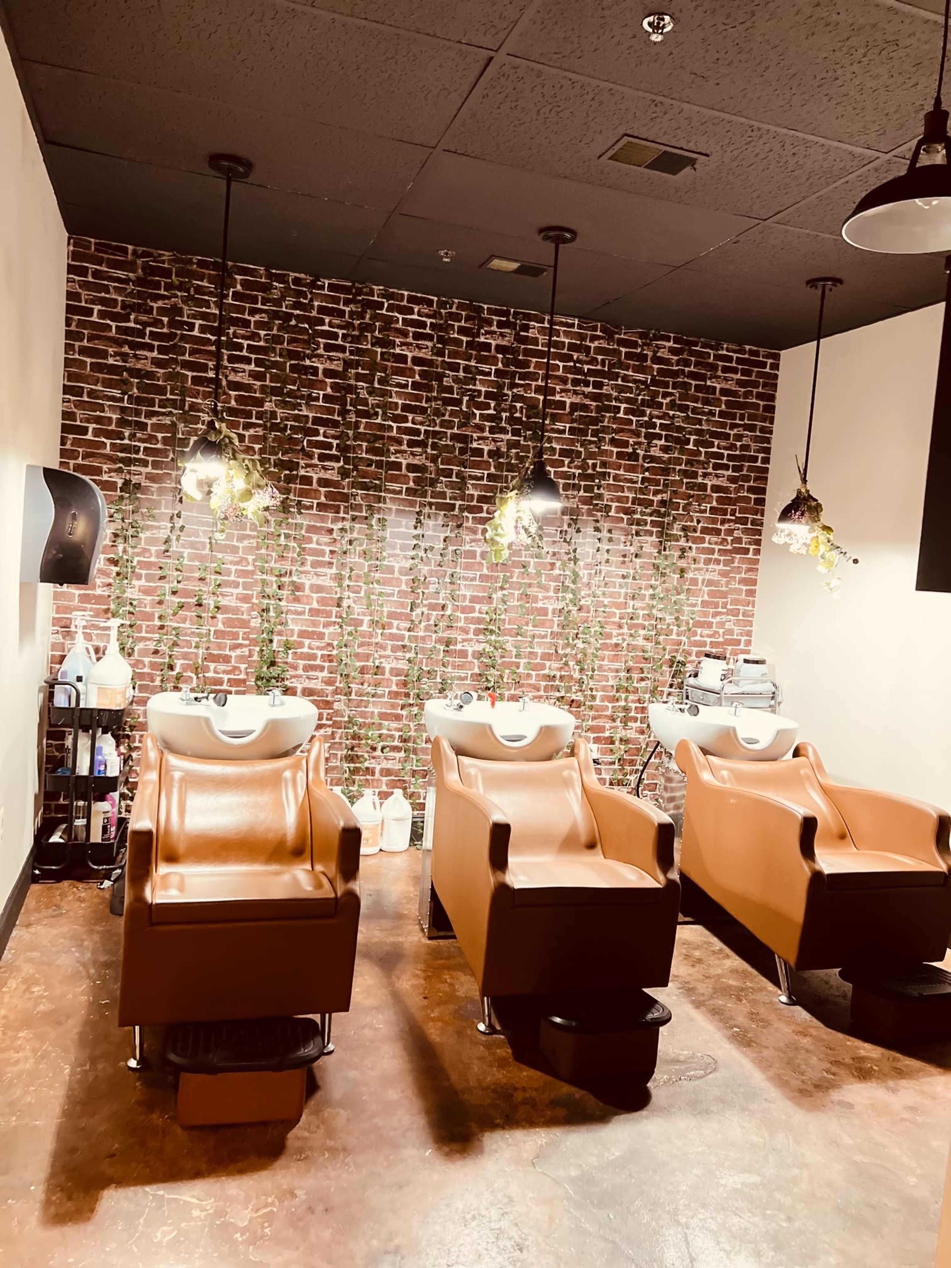 The image shows a hair salon area with three brown shampoo chairs arranged in front of a brick wall, adorned with hanging plants and pendant lights.