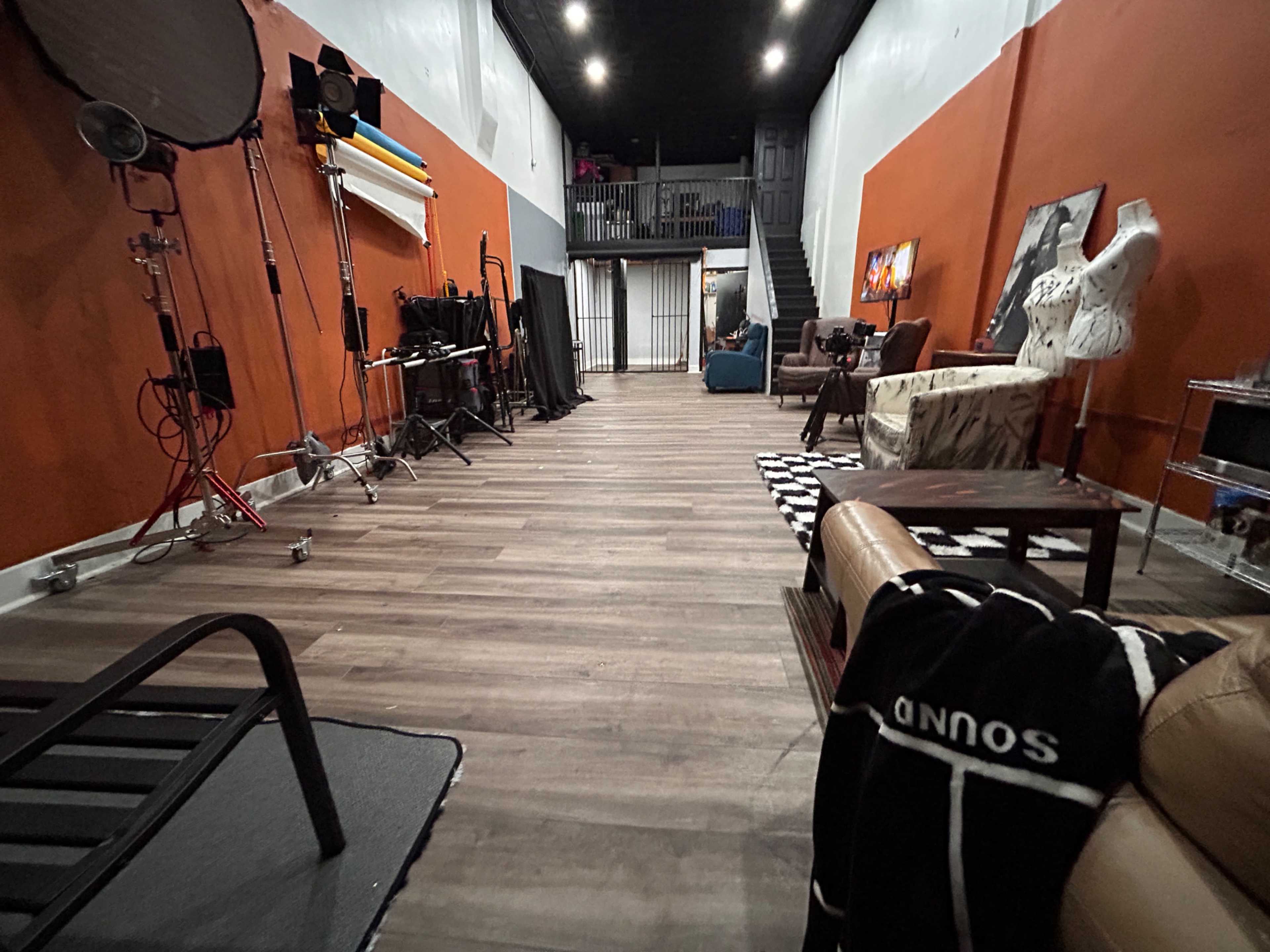 The image shows a spacious studio with orange walls, wooden flooring, and various props, including chairs, a mannequin, and lighting equipment.