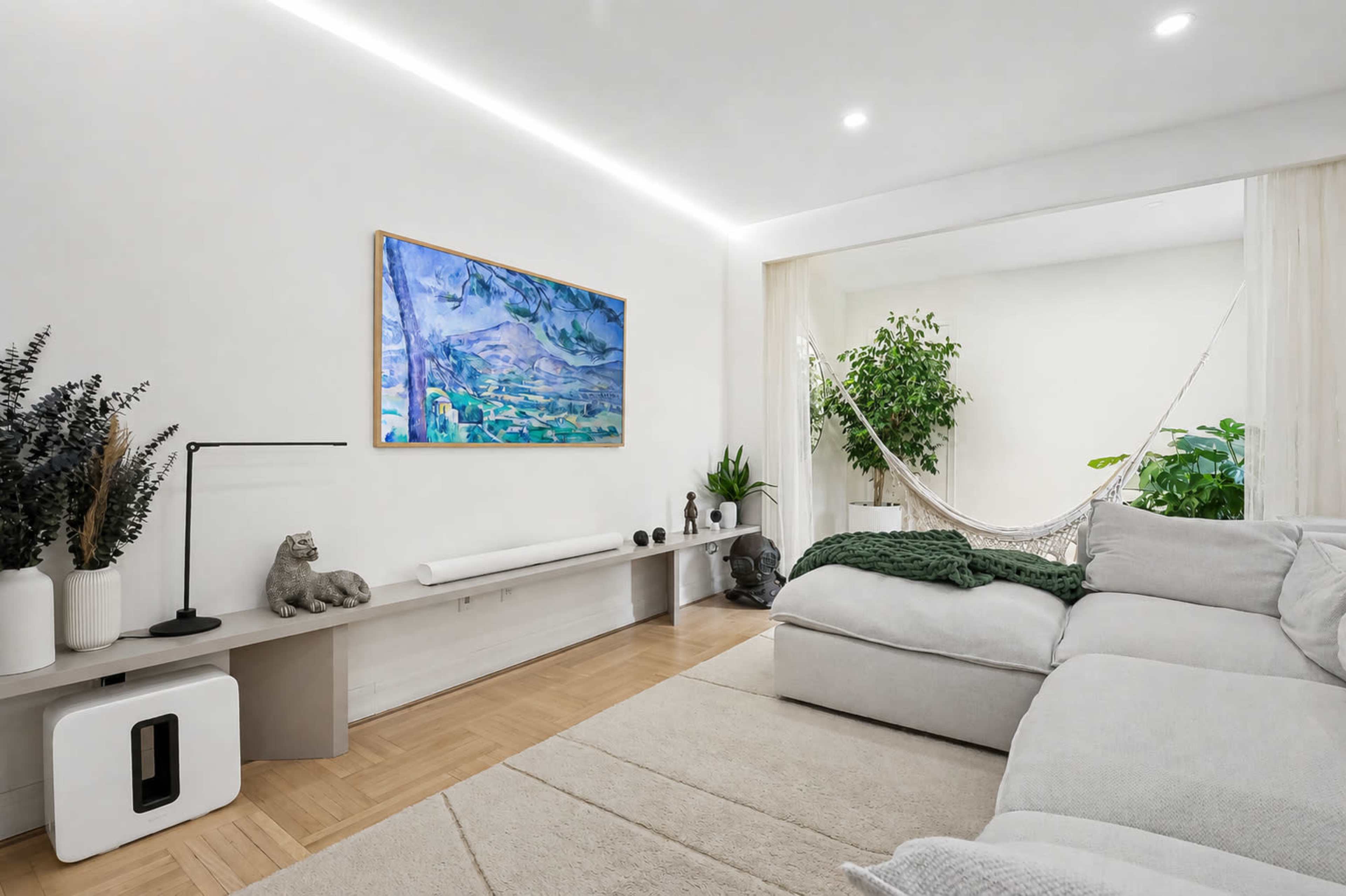 The image shows a modern living room featuring a large sectional sofa, a flat-screen TV, a decorative painting on the wall, and a hammock hanging in a small nook surrounded by plants.