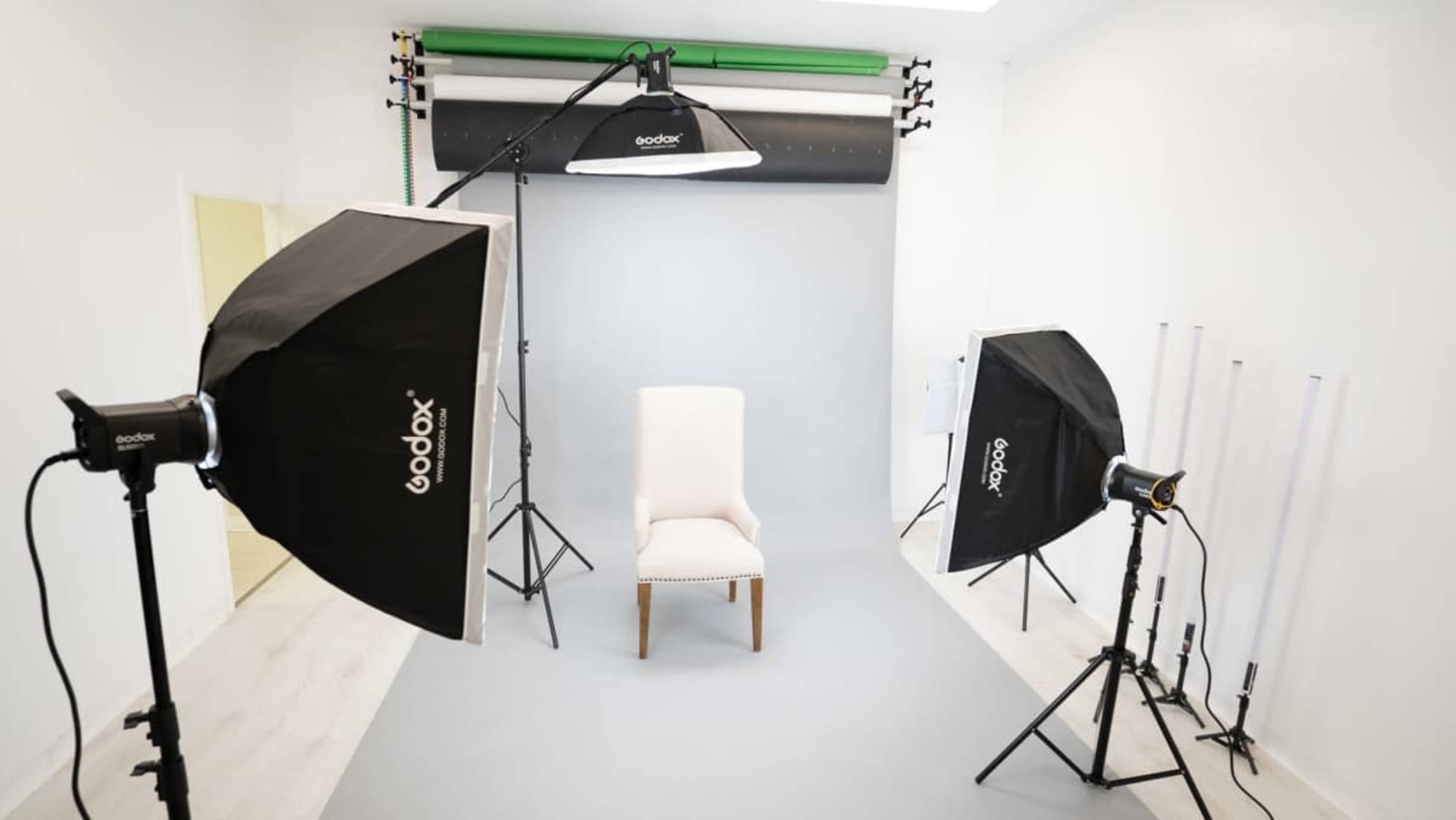 The image shows a photography studio setup featuring a white chair in front of a gray backdrop, flanked by two softbox lights and a spotlight.
