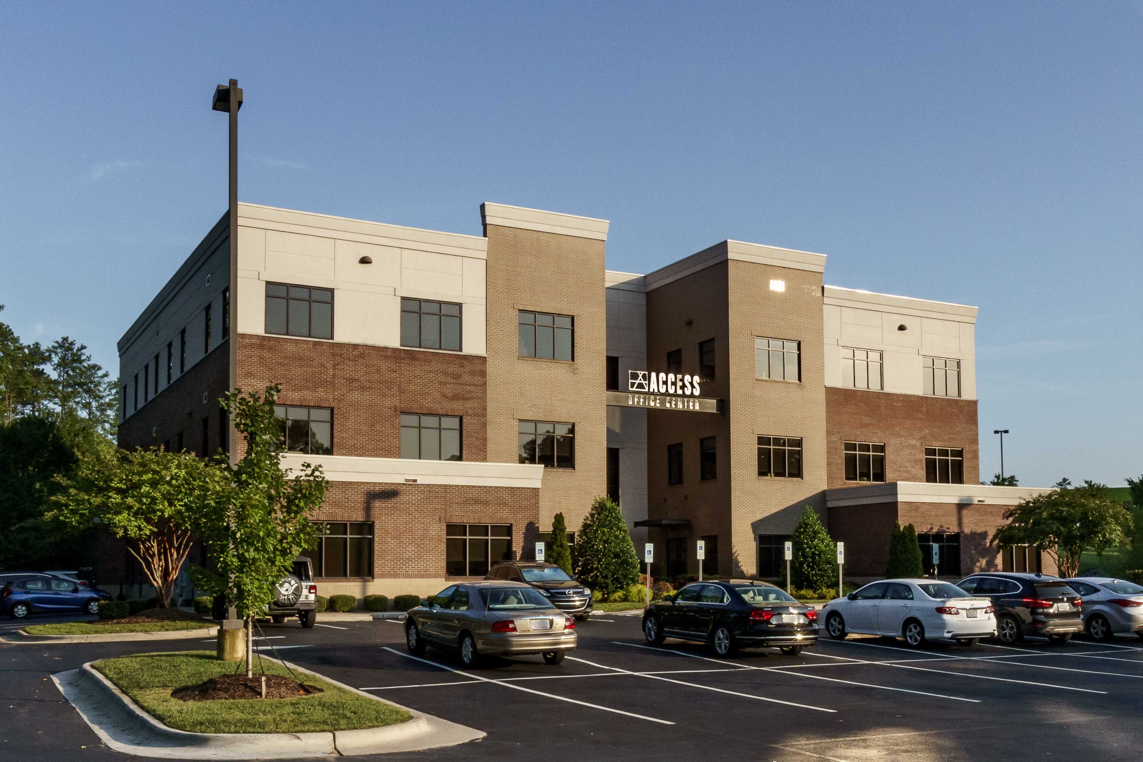 A modern, three-story brick office building with large windows and a parking lot filled with vehicles.