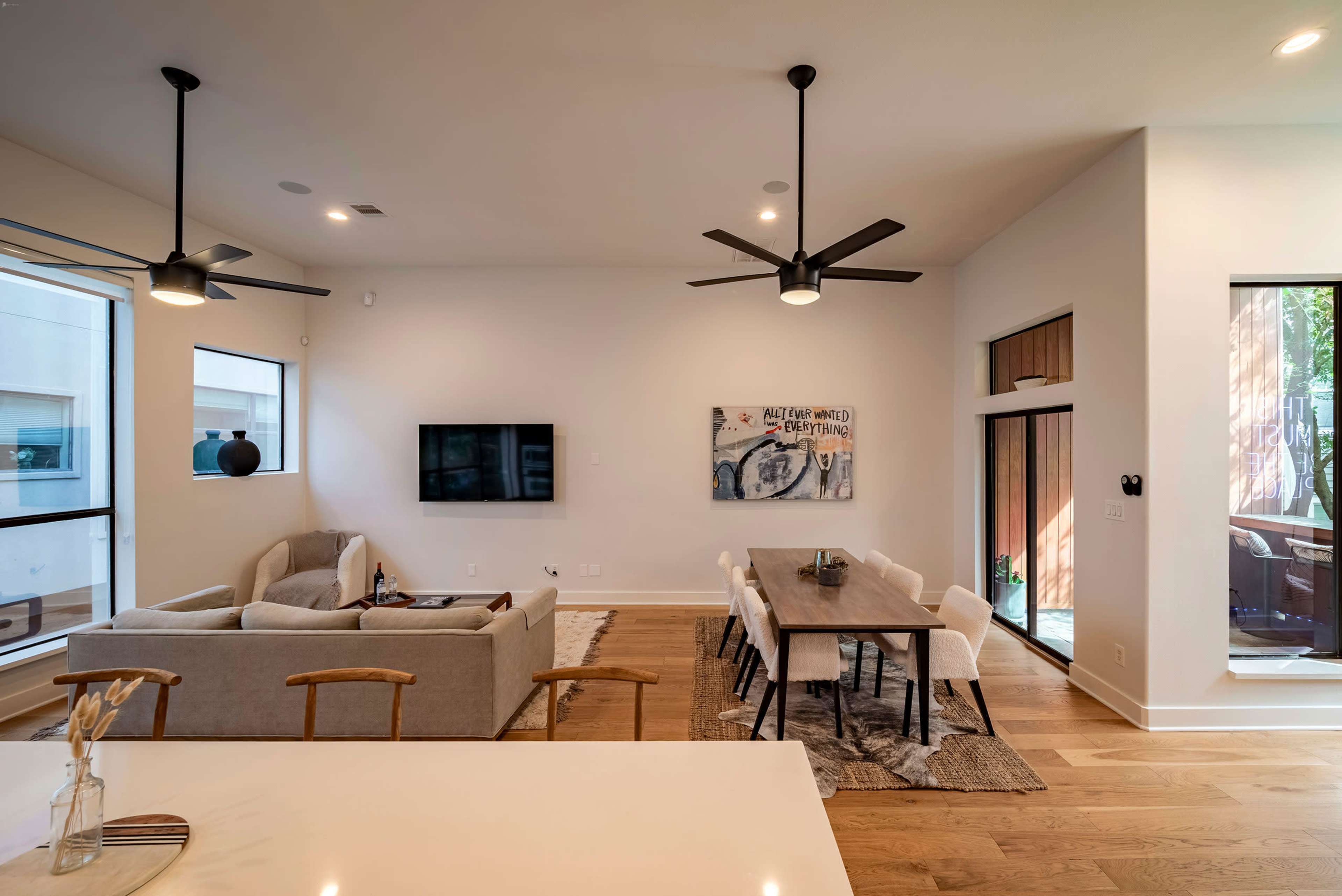 The image shows a modern living room with a gray sectional sofa and a dining table with chairs, featuring large windows and ceiling fans.