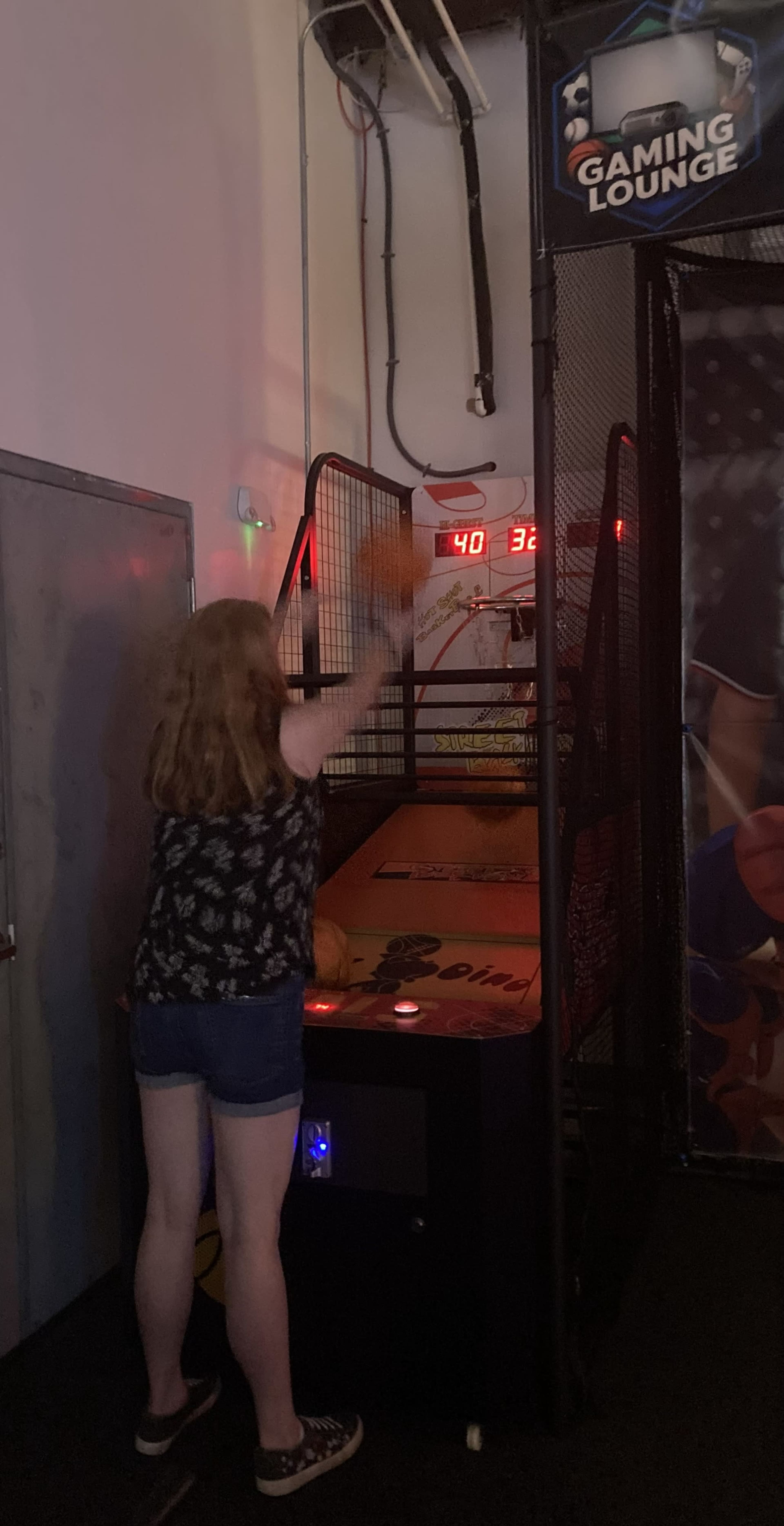 A person is playing a basketball arcade game in a gaming lounge, with scores displayed on the machine's screen.