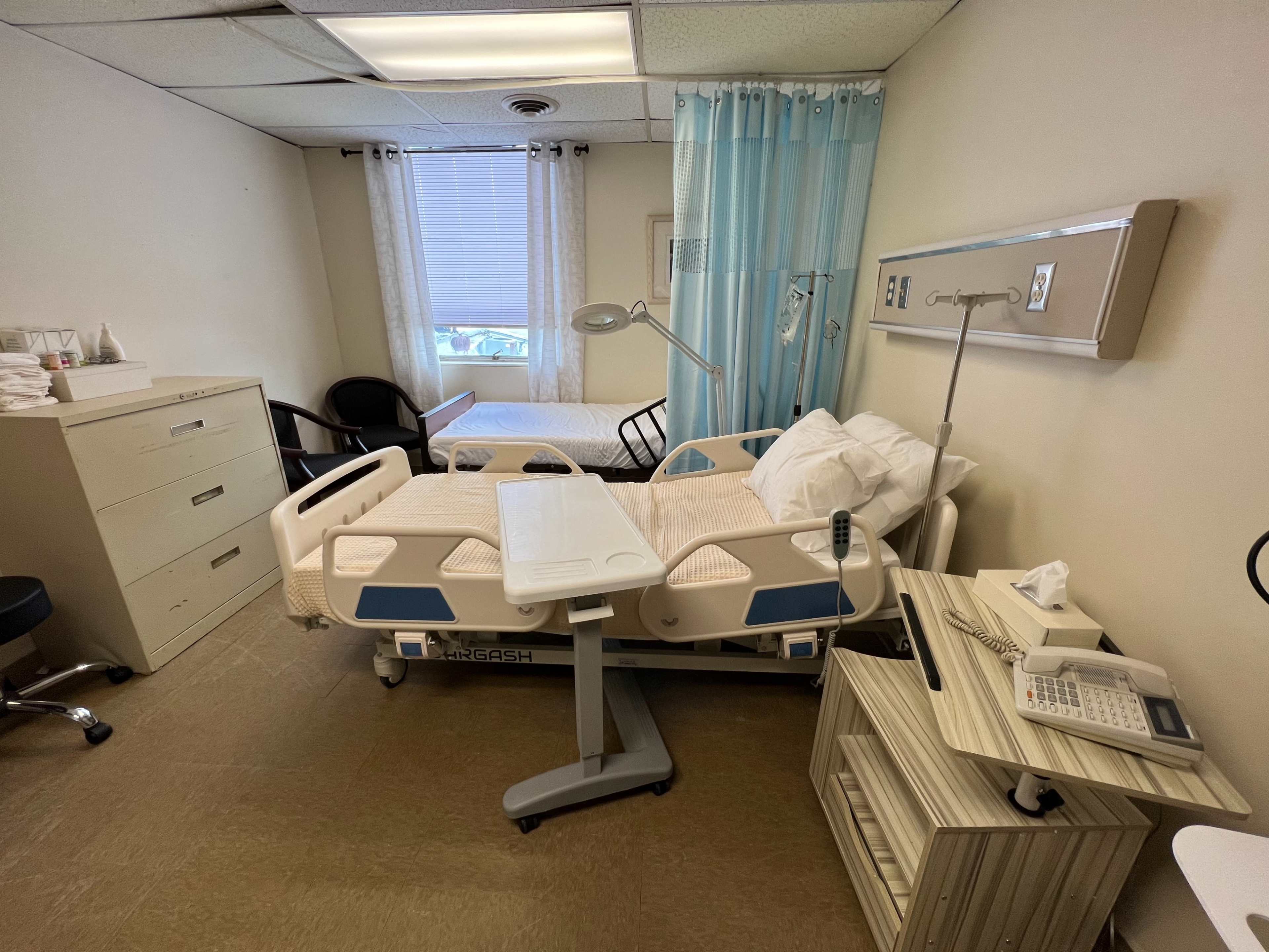 The image shows a hospital room with a bed, a chair, a bedside table, a desk, and a window.