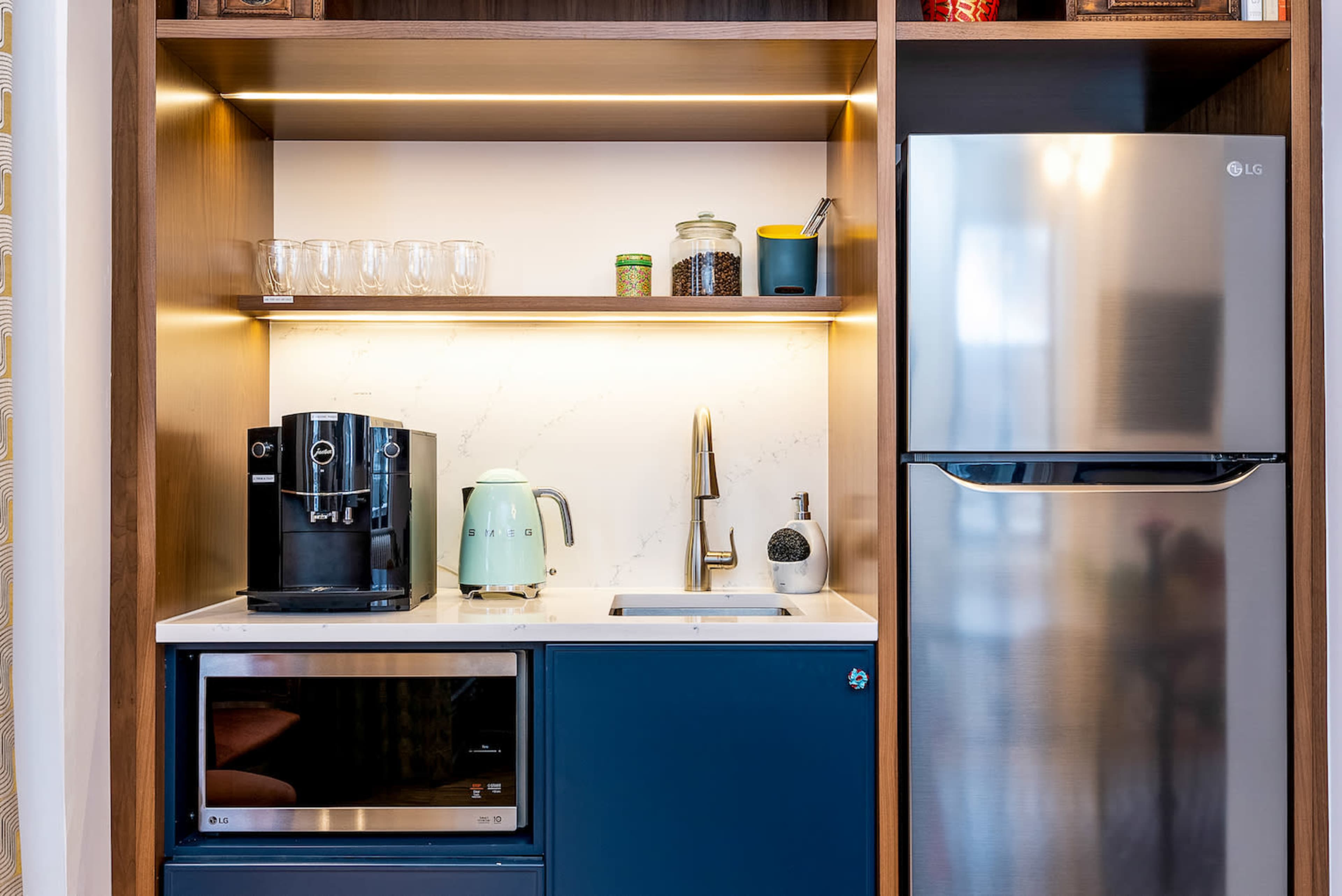 A modern kitchenette featuring a coffee maker, kettle, sink, and refrigerator, all arranged within wooden cabinetry with illuminated shelves.