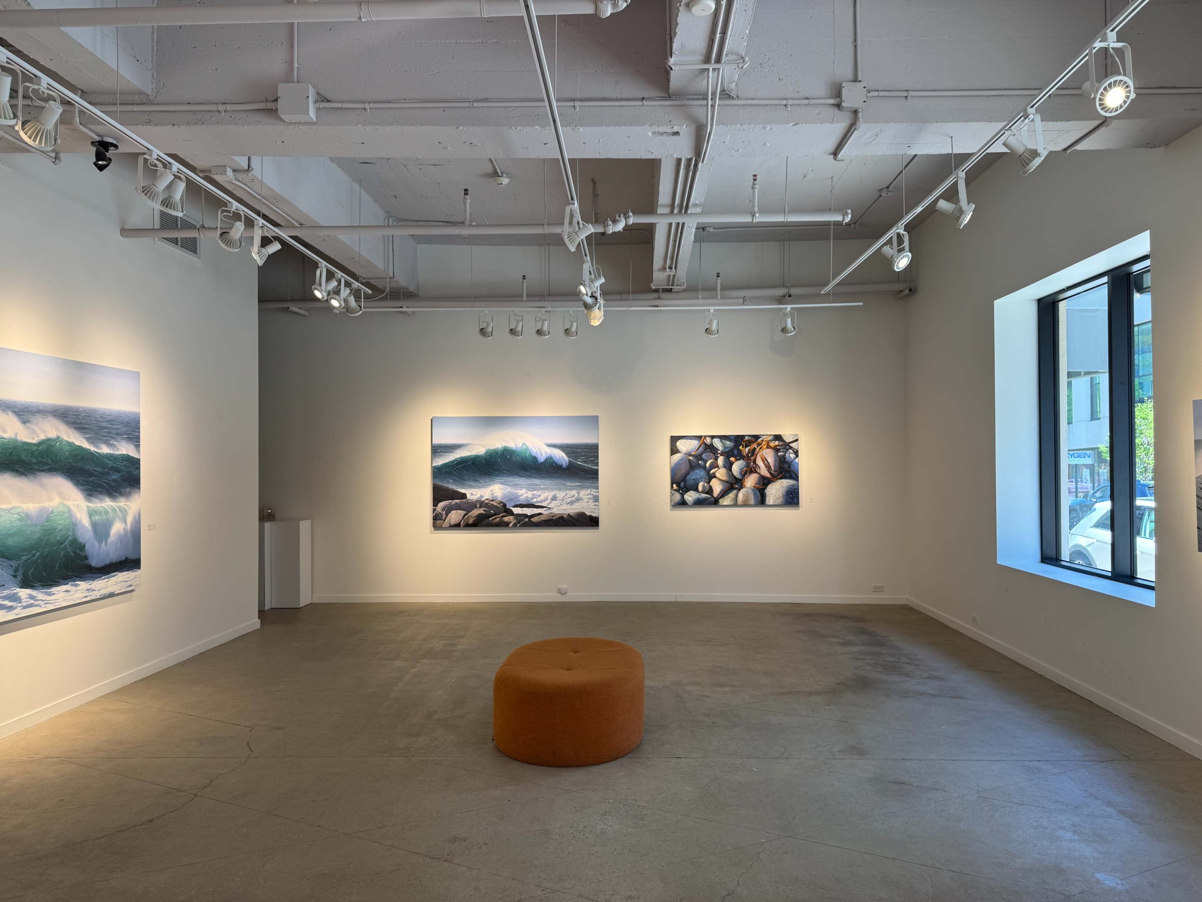 The image shows an art gallery with large ocean-themed photographs on the walls and an orange circular ottoman in the center of the room.