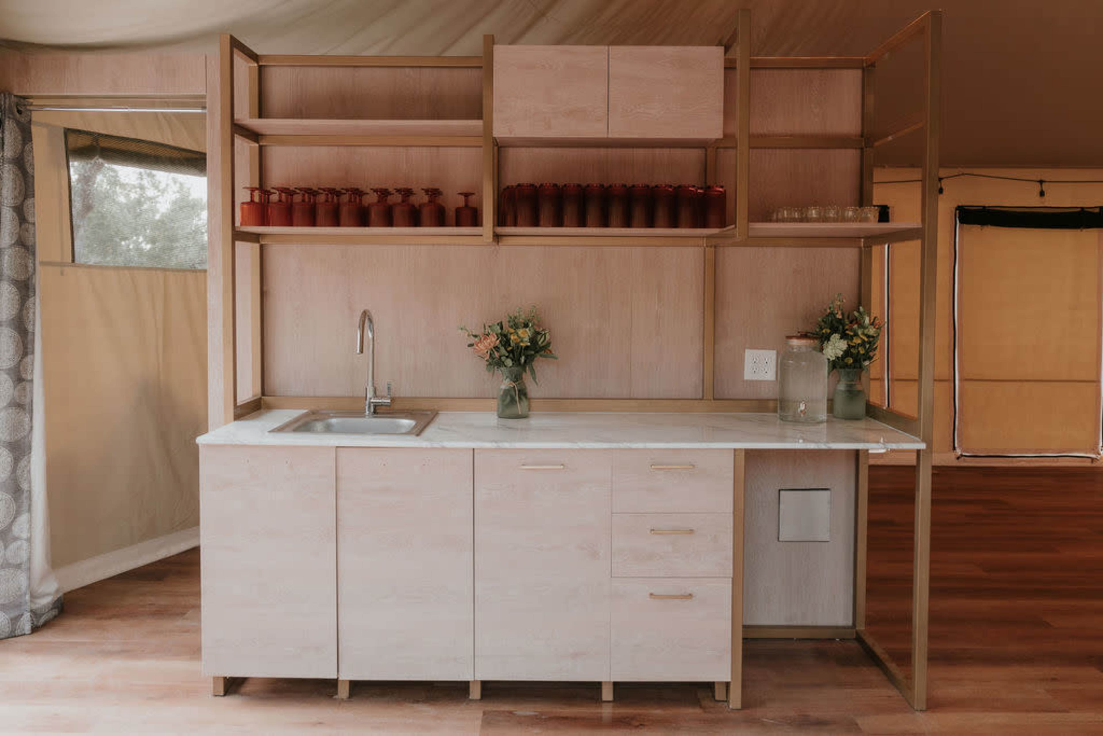 The image shows a modern kitchen space with a minimalist design, featuring a sink, cabinets, and open shelving with glassware and flowers.