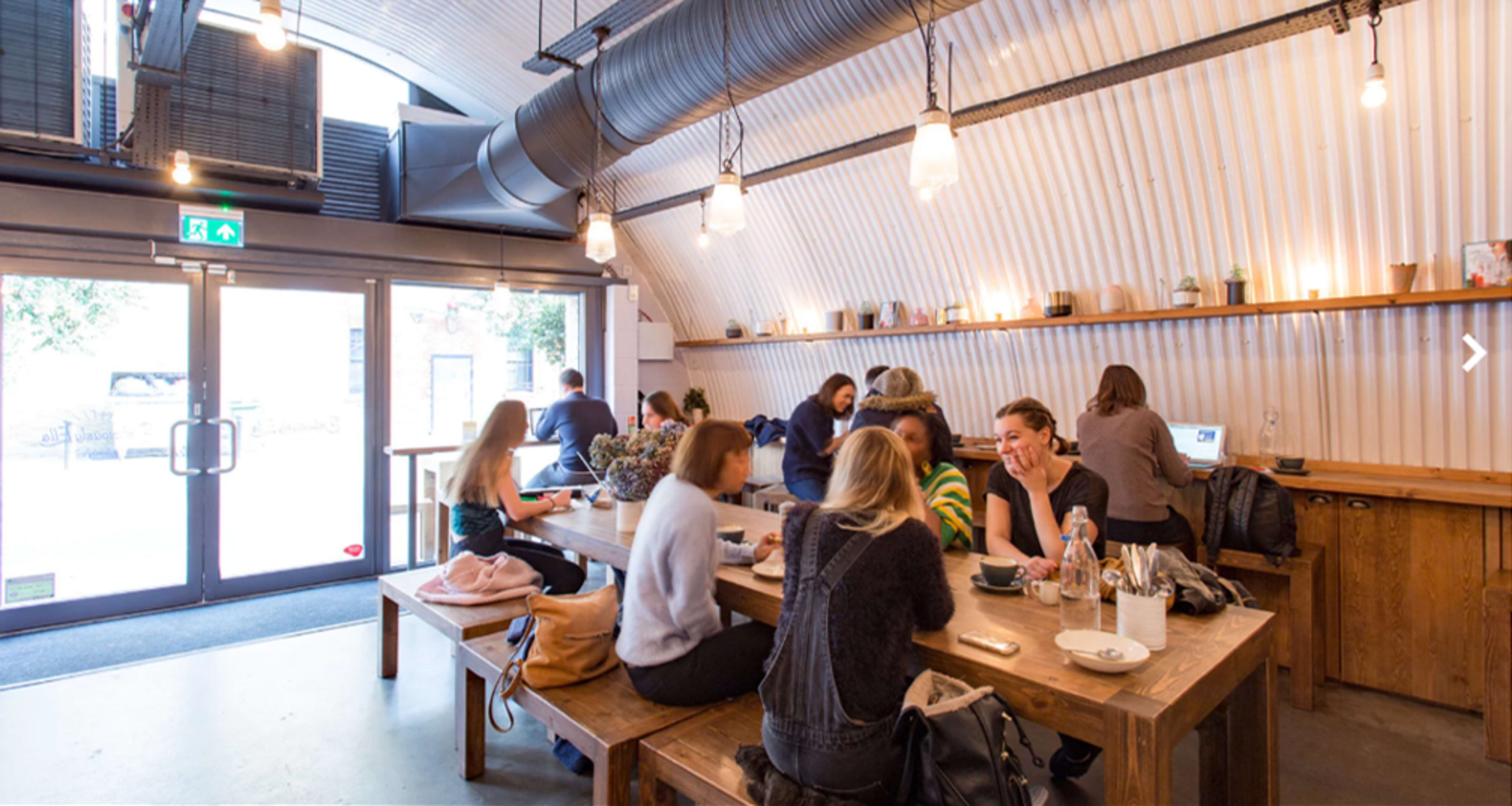 A group of people are seated at wooden tables in a spacious, well-lit cafe with large windows and industrial-style decor.