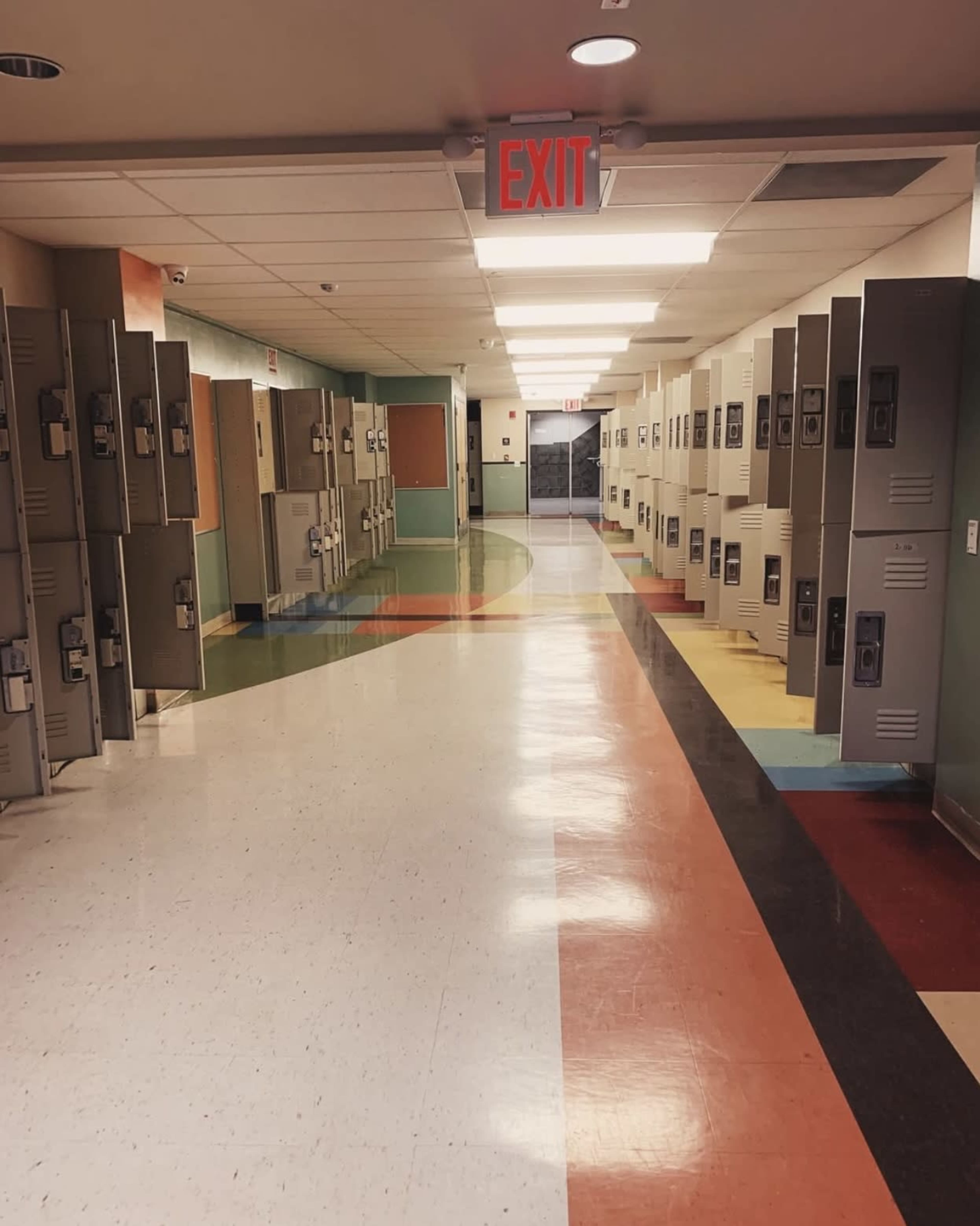 The image shows a brightly colored school hallway lined with lockers on both sides and an exit sign at the far end.
