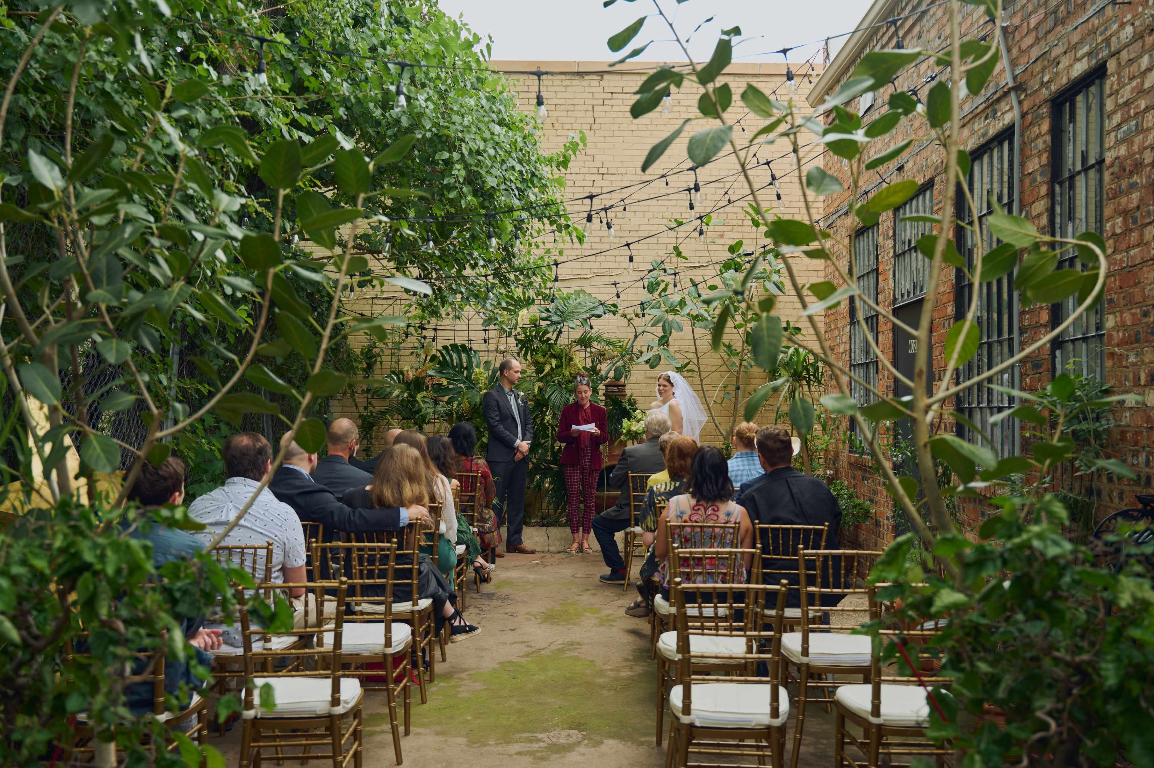 A wedding ceremony takes place in a lush, green courtyard surrounded by plants, with guests seated on gold chiavari chairs.