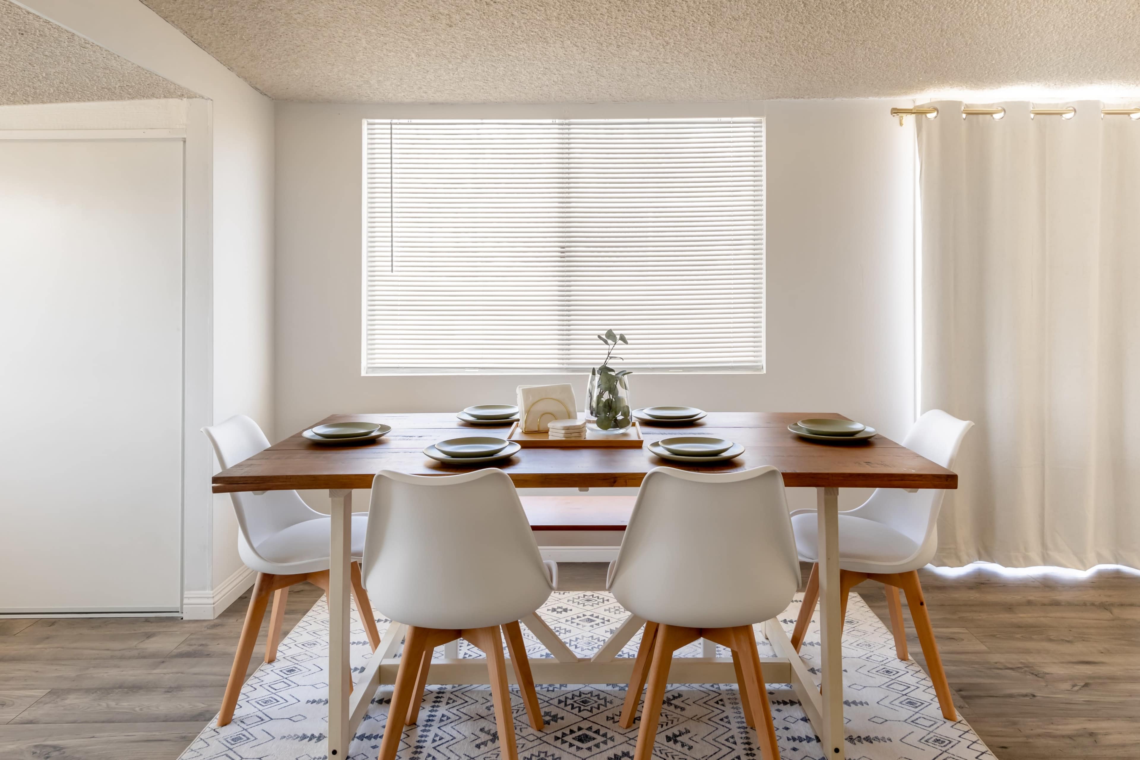 A wooden dining table is set with dishes and surrounded by four white chairs in a well-lit room with a window and a curtain.